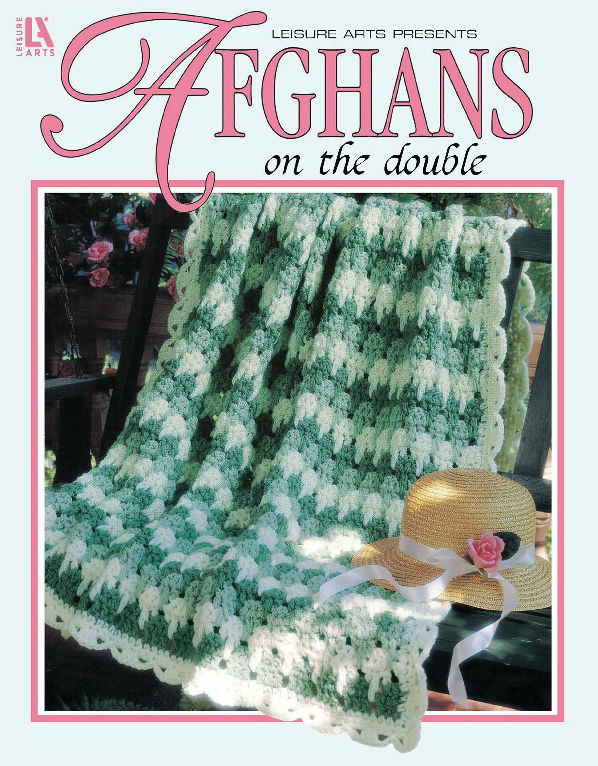 A green and white crocheted afghan with scalloped edges from Leisure Arts’ Afghans on the Double Digital Download is draped over a wooden chair. Nearby, a straw hat with a pink flower and blue ribbon rests.