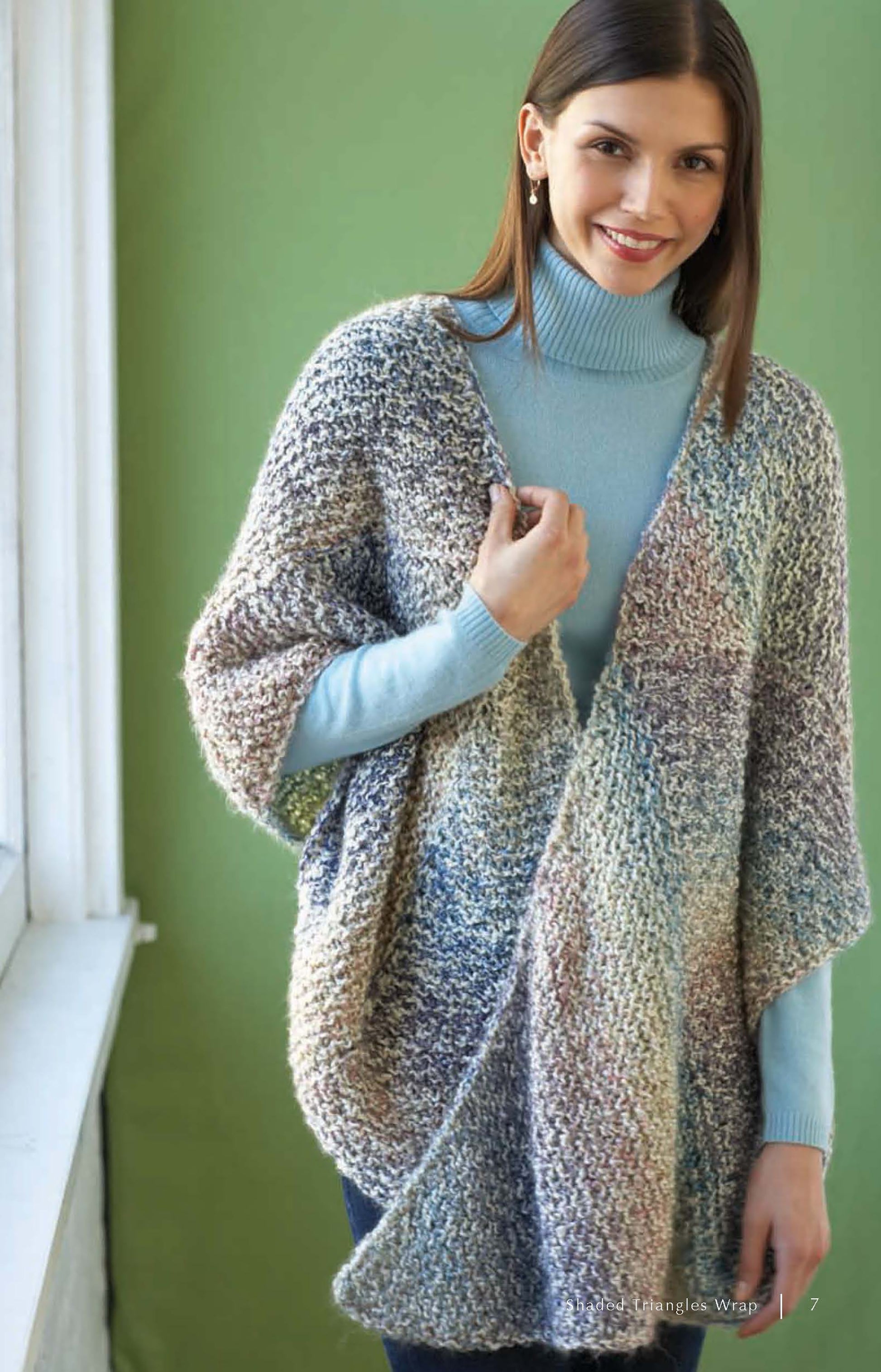 A woman in a light blue turtleneck and a chunky, colorful wrap made from bulky yarn stands by a window—perfect inspiration for scarves and shawls featured in Cozy Cover-Ups by Leisure Arts, with 4 crochet & 2 knit designs using Lion Brand Homespun.