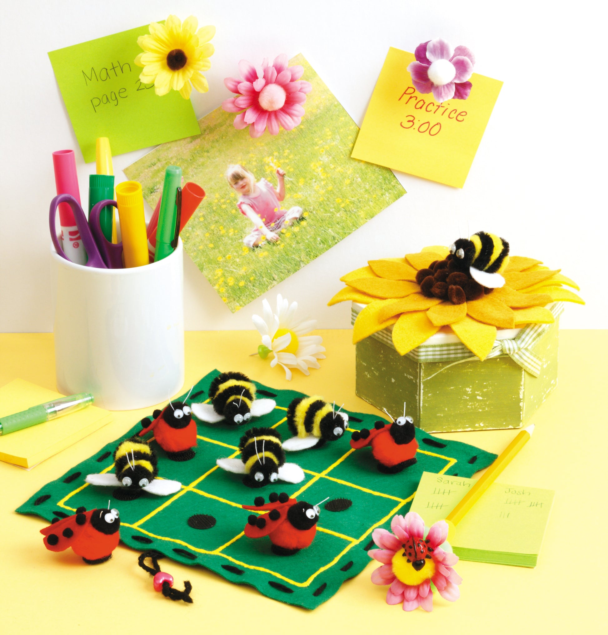 A cheerful desk shows Leisure Arts Pompom Crafts: pens in a cup, sticky notes, a sunflower box with bee toys, a felt game board with bee and ladybug pieces for flower and animal crafts, plus a photo of a child among daisies.