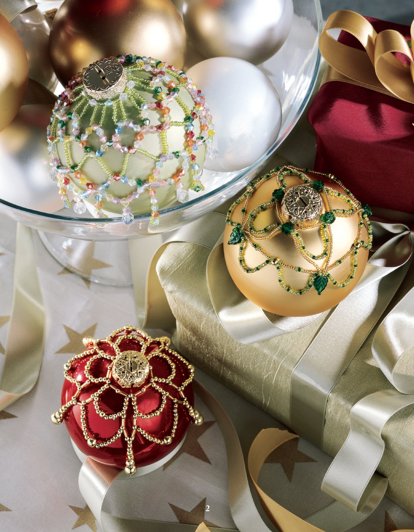 A glass bowl displays gold, white, and green Christmas ornaments with detailed patterns from Leisure Arts' Big Book of Beaded Ornaments. Red and gold beaded ornaments are arranged beside a gold-wrapped gift and a red box with a gold ribbon.