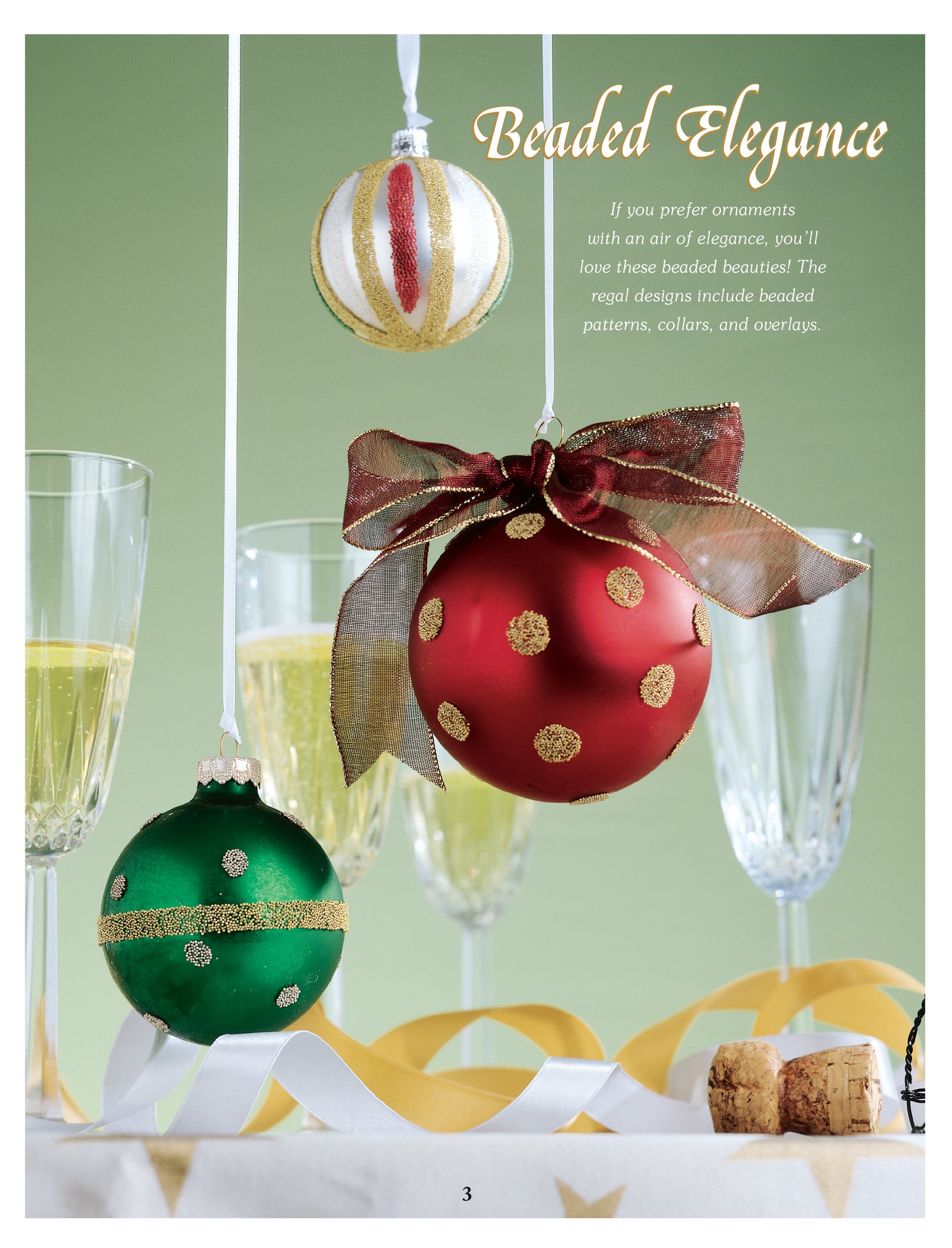 Three beaded Christmas ornaments from Leisure Arts’ Big Book of Beaded Ornaments—red with gold polka dots, green with gold accents, and white with stripes—are displayed in front of champagne glasses on a festive table.