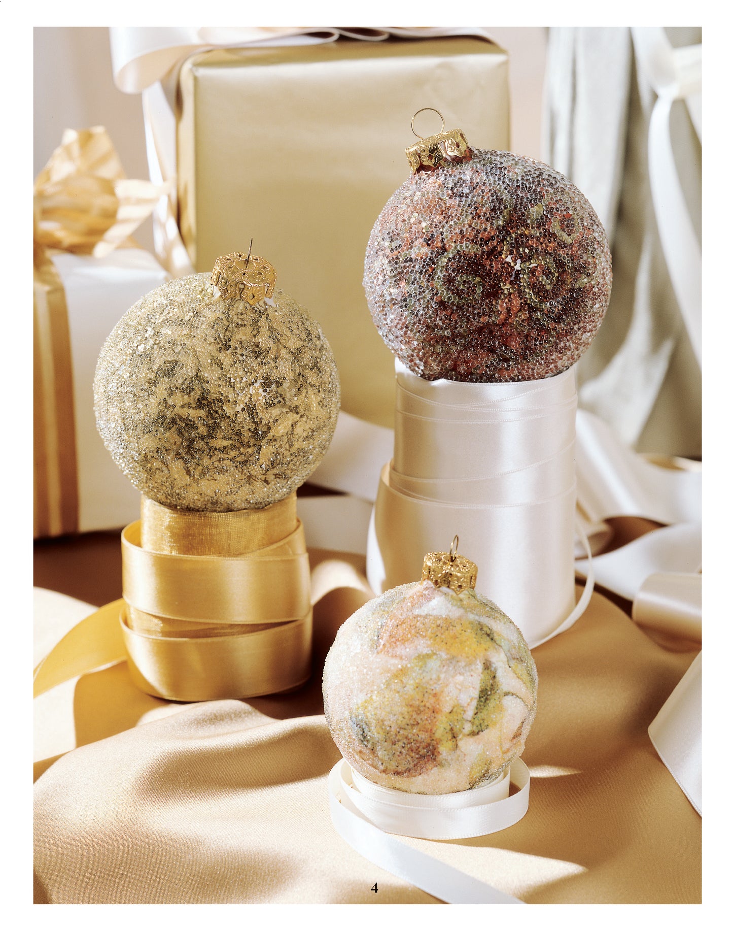 Three beaded Christmas ornaments made with patterns from Leisure Arts' Big Book of Beaded Ornaments hang on silk ribbon amid gold and white wrapped gifts. Soft lighting adds an elegant, festive holiday touch.