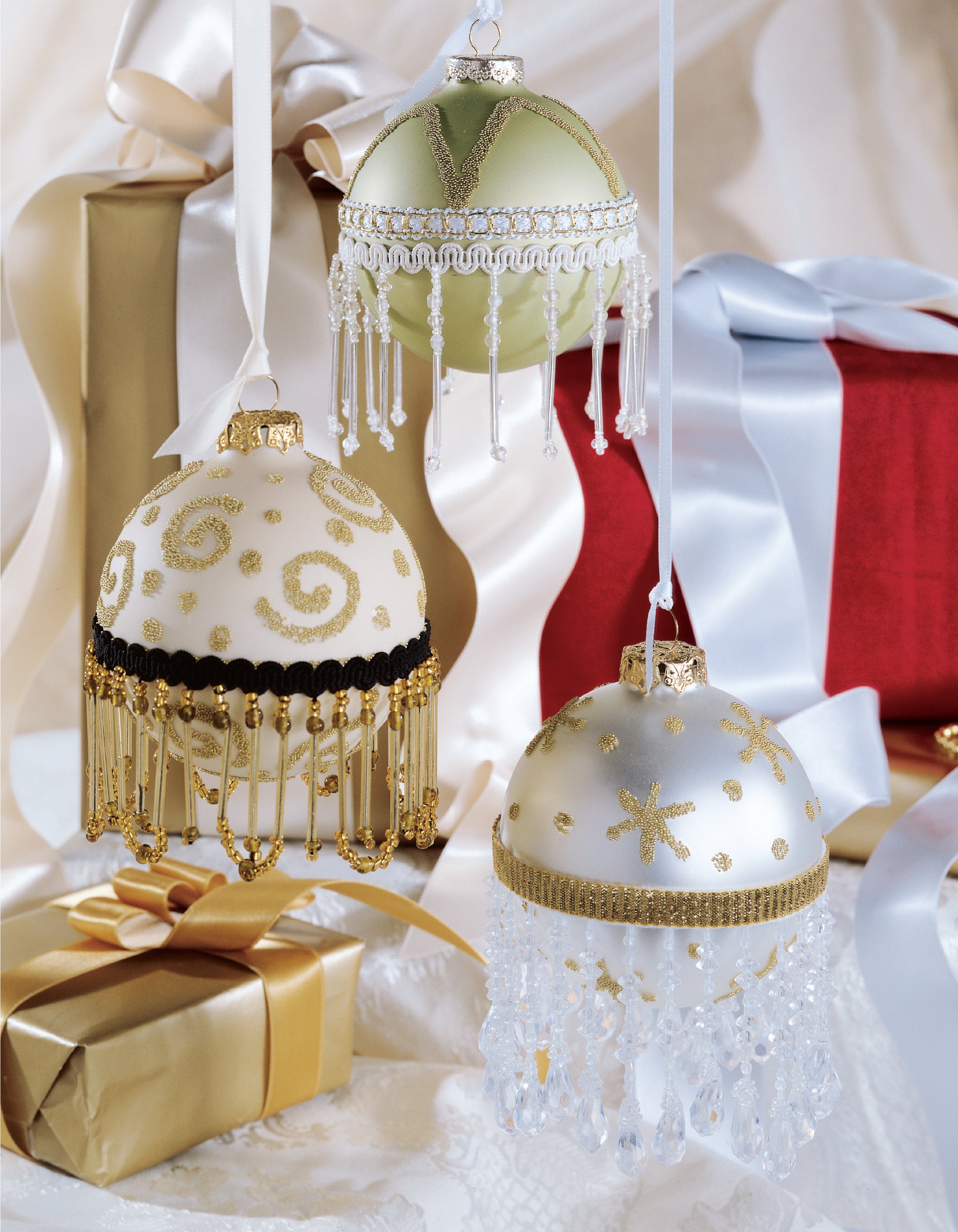 Three beaded Christmas ornaments with gold and silver details hang before red and gold gift boxes, inspired by the "Big Book of Beaded Ornaments" from Leisure Arts—a perfect guide for crafting beautiful holiday decorations.