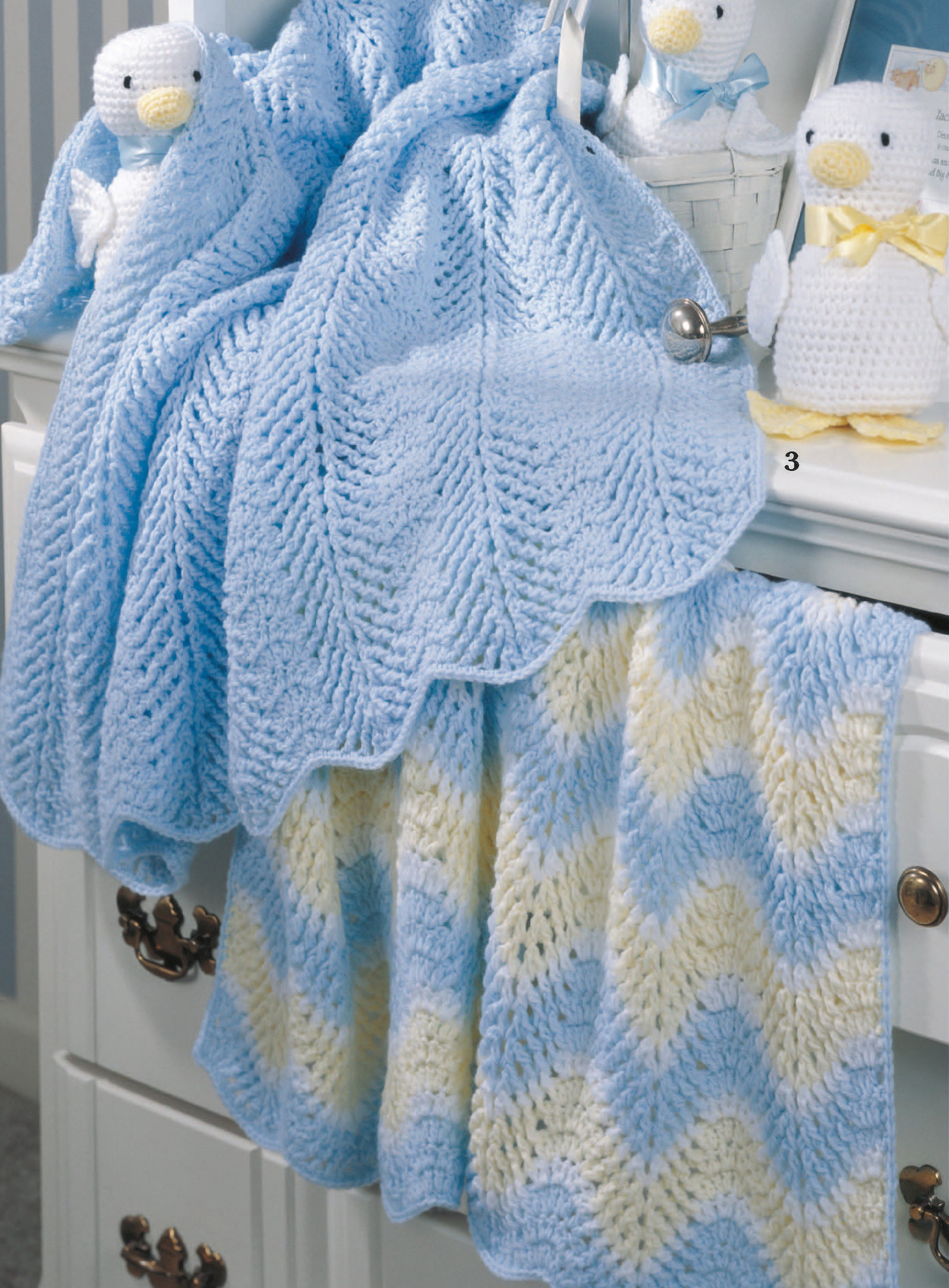 A soft blue Just Ducky Baby Afghan by Leisure Arts drapes over a dresser. Below, a striped knit afghan hangs while two crocheted ducks with yellow ribbons sit on top—adorable nursery gifts from Leisure Arts.
