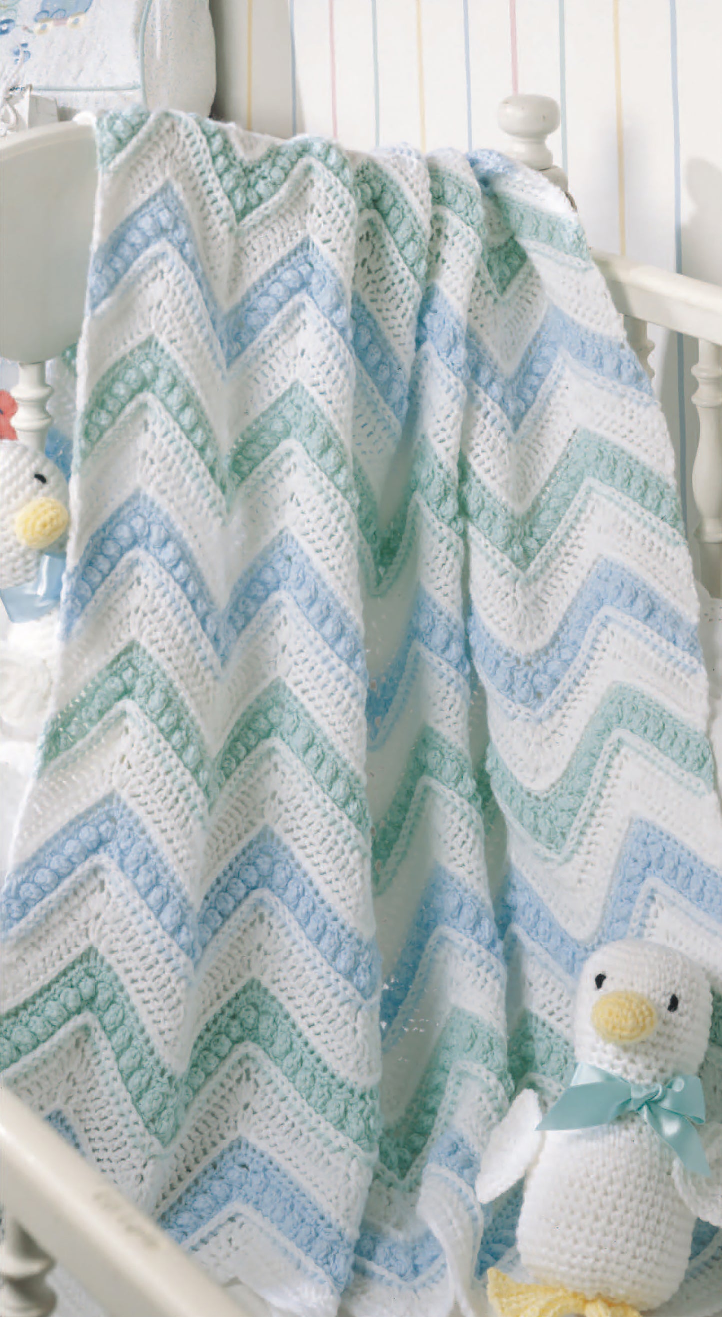 The Just Ducky Baby Afghans by Leisure Arts, featuring a zigzag pattern in white, blue, and green, drapes over the crib—an ideal nursery gift. A plush duck toy with a blue ribbon rests nearby against pastel-striped wallpaper.