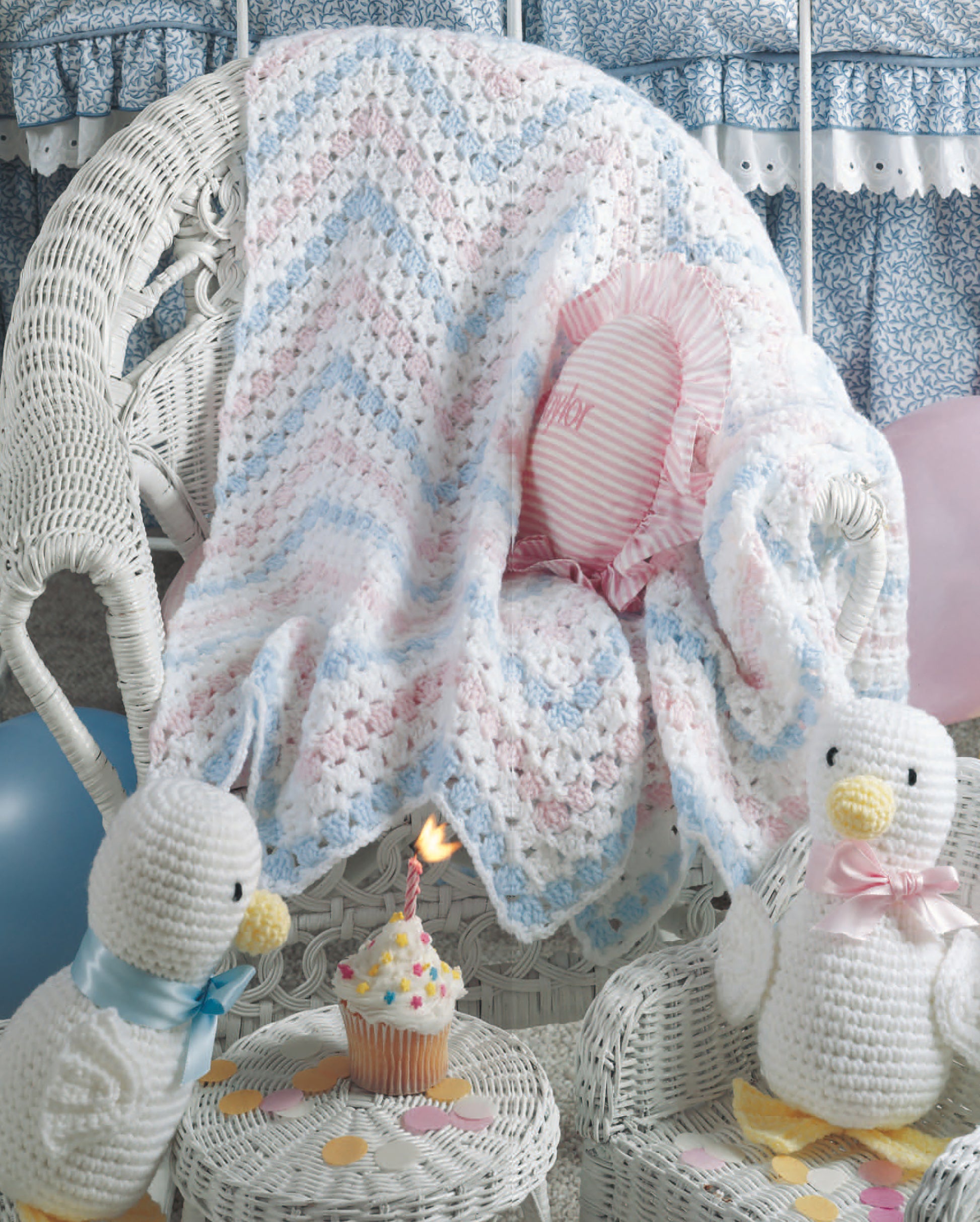 Just Ducky Baby Afghans by Leisure Arts drape over a white wicker chair, surrounded by crochet ducks, pastel balloons, and a cupcake with a lit candle—perfect for charming nursery gifts.