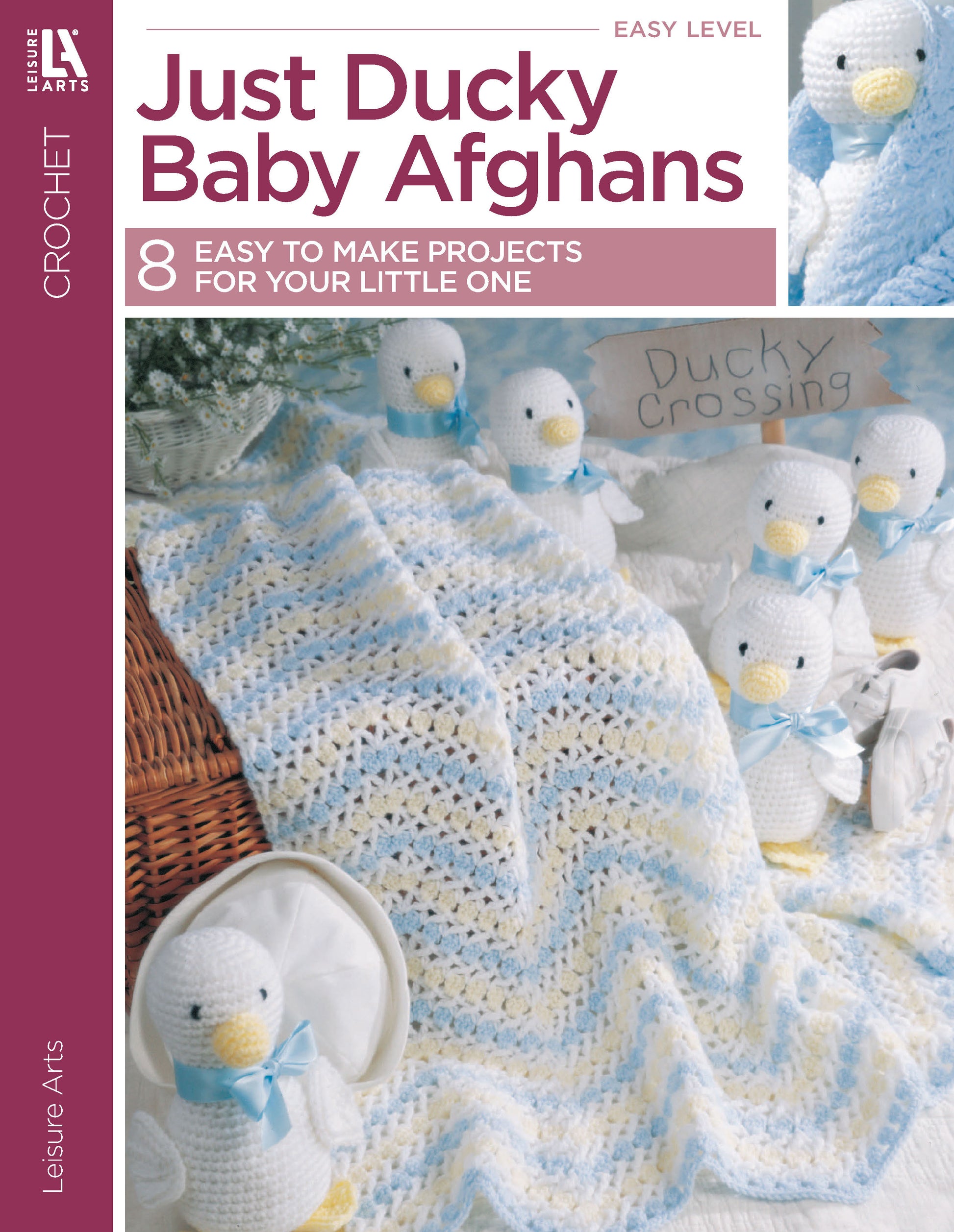 The Leisure Arts Just Ducky Baby Afghans are draped over a chair, surrounded by crocheted white duck toys with yellow beaks and blue ribbons—a charming nursery gift. A "Ducky Crossing" sign appears in the background.