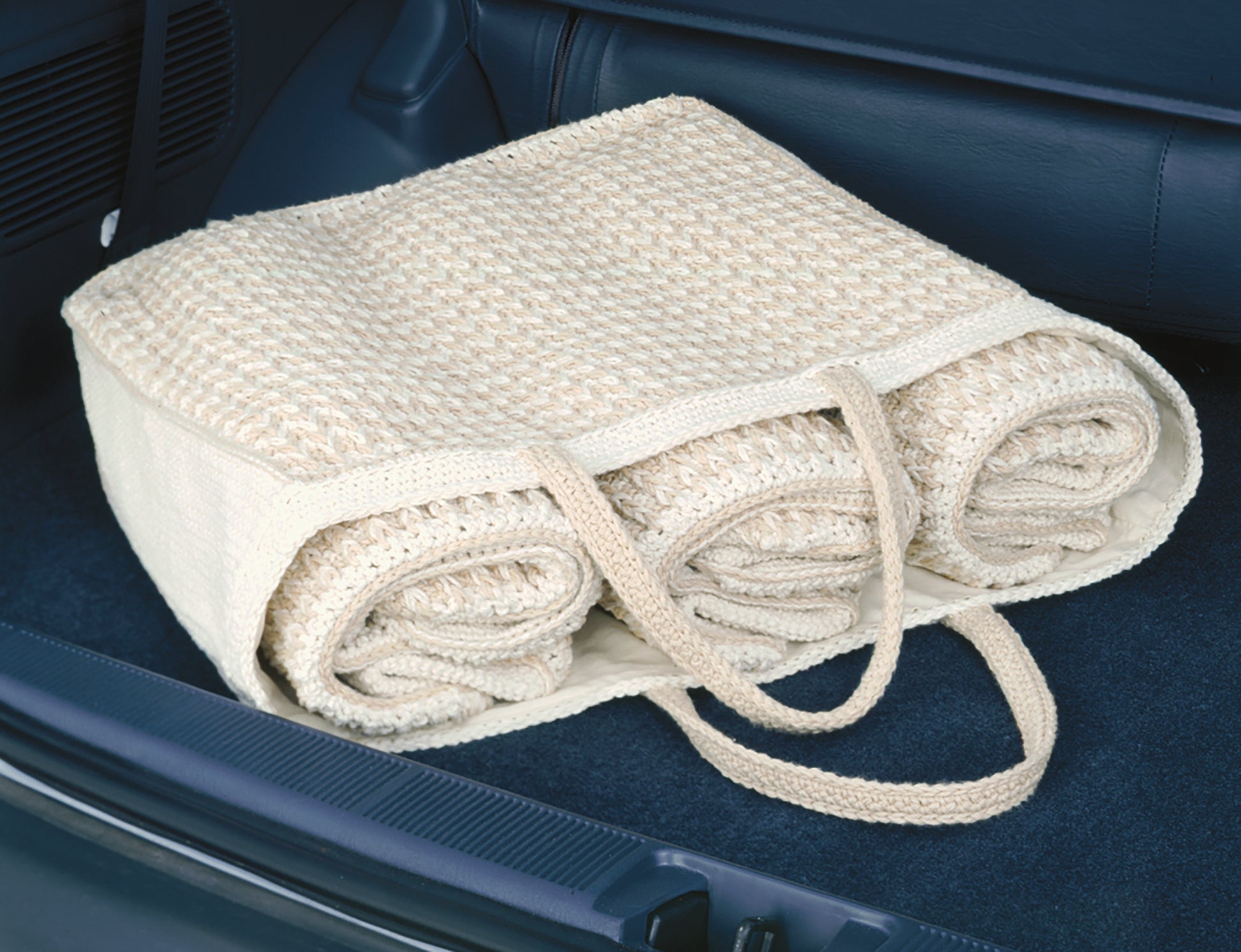 The Enviro-Totes Digital Download by Leisure Arts features a beige woven bag, inspired by crochet tote patterns, holding three rolled-up knit blankets on the back seat of a car with dark blue upholstery.