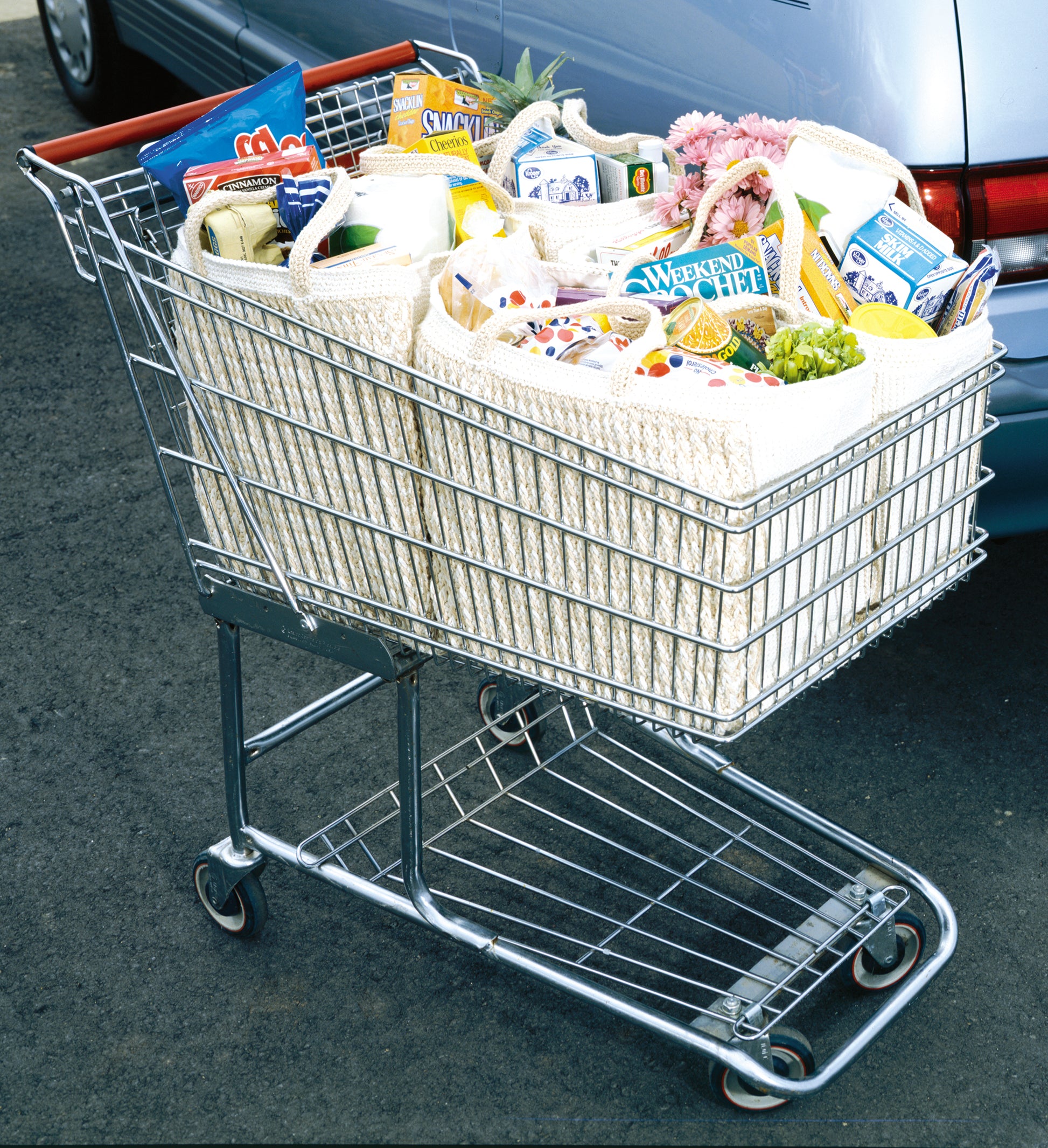 A shopping cart with groceries—boxed food, produce, and flowers—packed in Leisure Arts' Enviro-Totes Digital Download reusable crochet bags stands on asphalt next to a parked car.