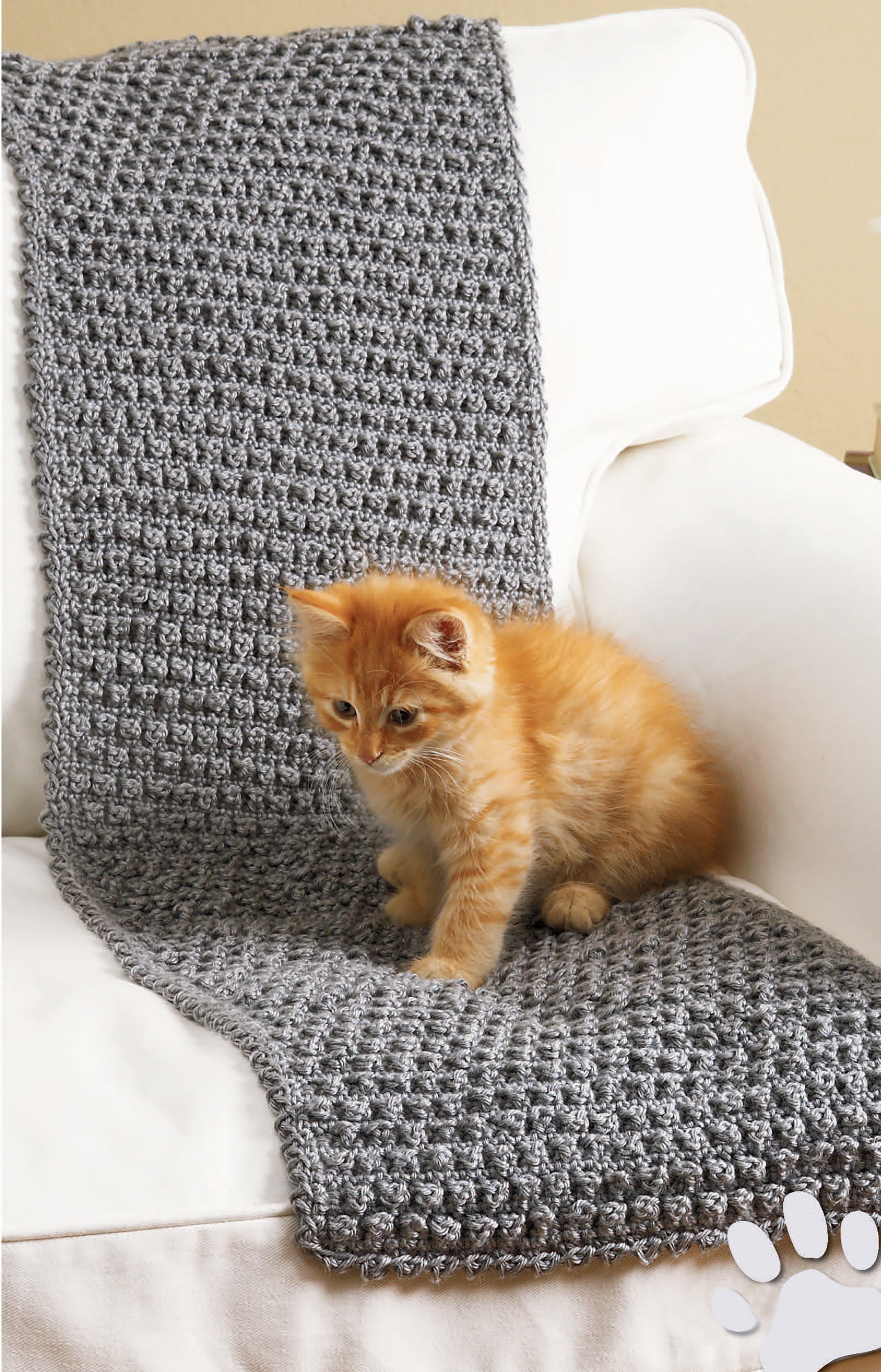 A fluffy orange kitten sits on a white chair with a textured gray knitted blanket, next to the Sweet Pet Comforts Digital Download pet bed by Leisure Arts, looking curiously downward.