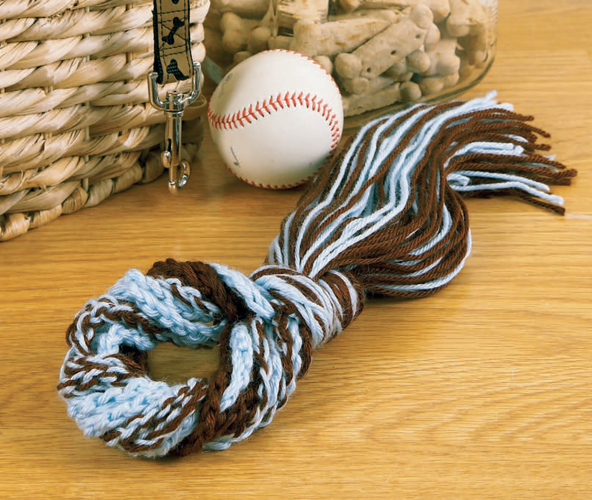 The Sweet Pet Comforts Digital Download by Leisure Arts features a twisted blue and brown dog toy made from medium weight yarn, shown on wood near a basket, baseball, treat jar, and a paw print leash.