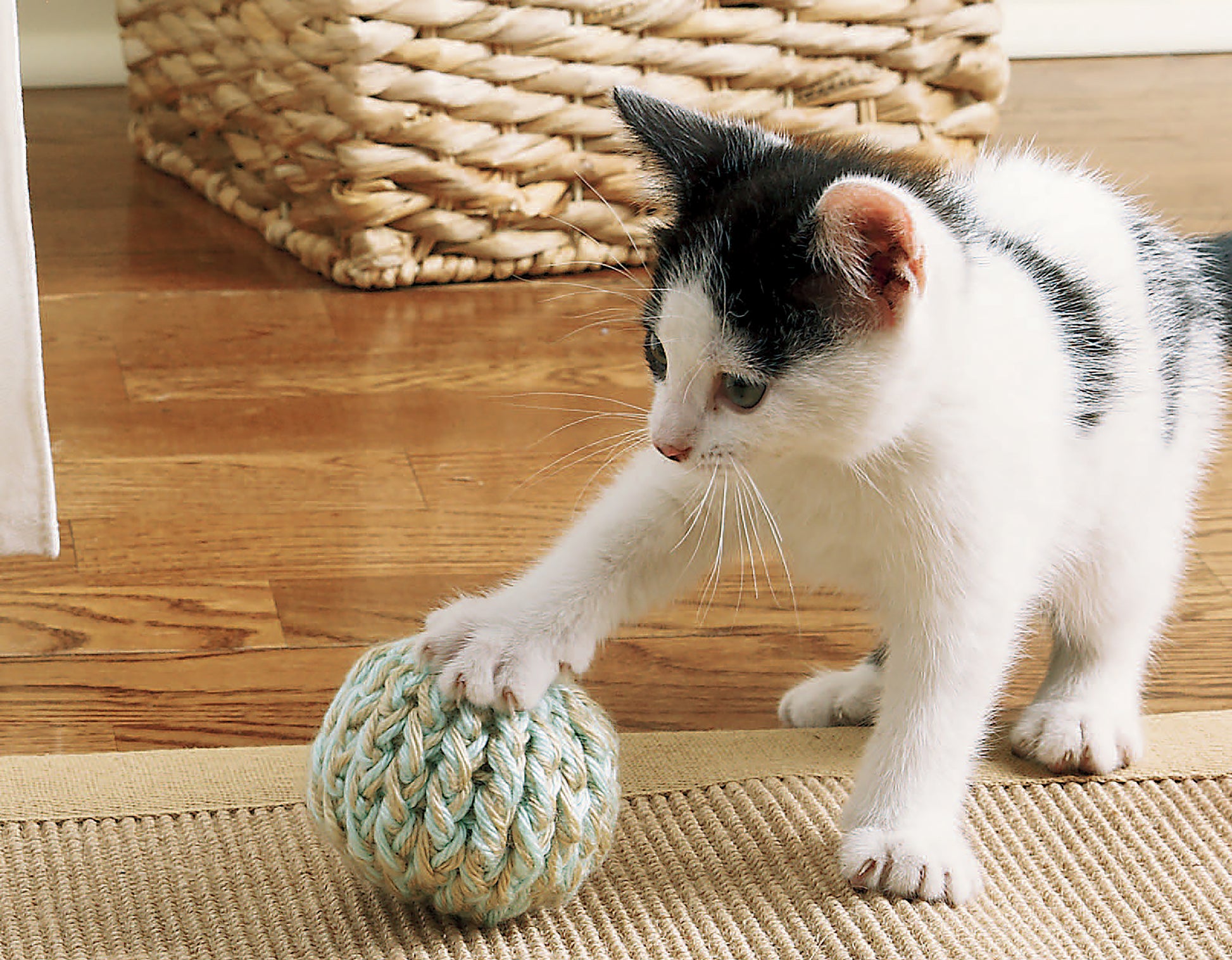 A black and white kitten plays with a yarn ball on a rug, while a cozy crochet bed made from medium weight yarn—pattern from Sweet Pet Comforts Digital Download by Leisure Arts—sits beside a woven basket on the wooden floor.