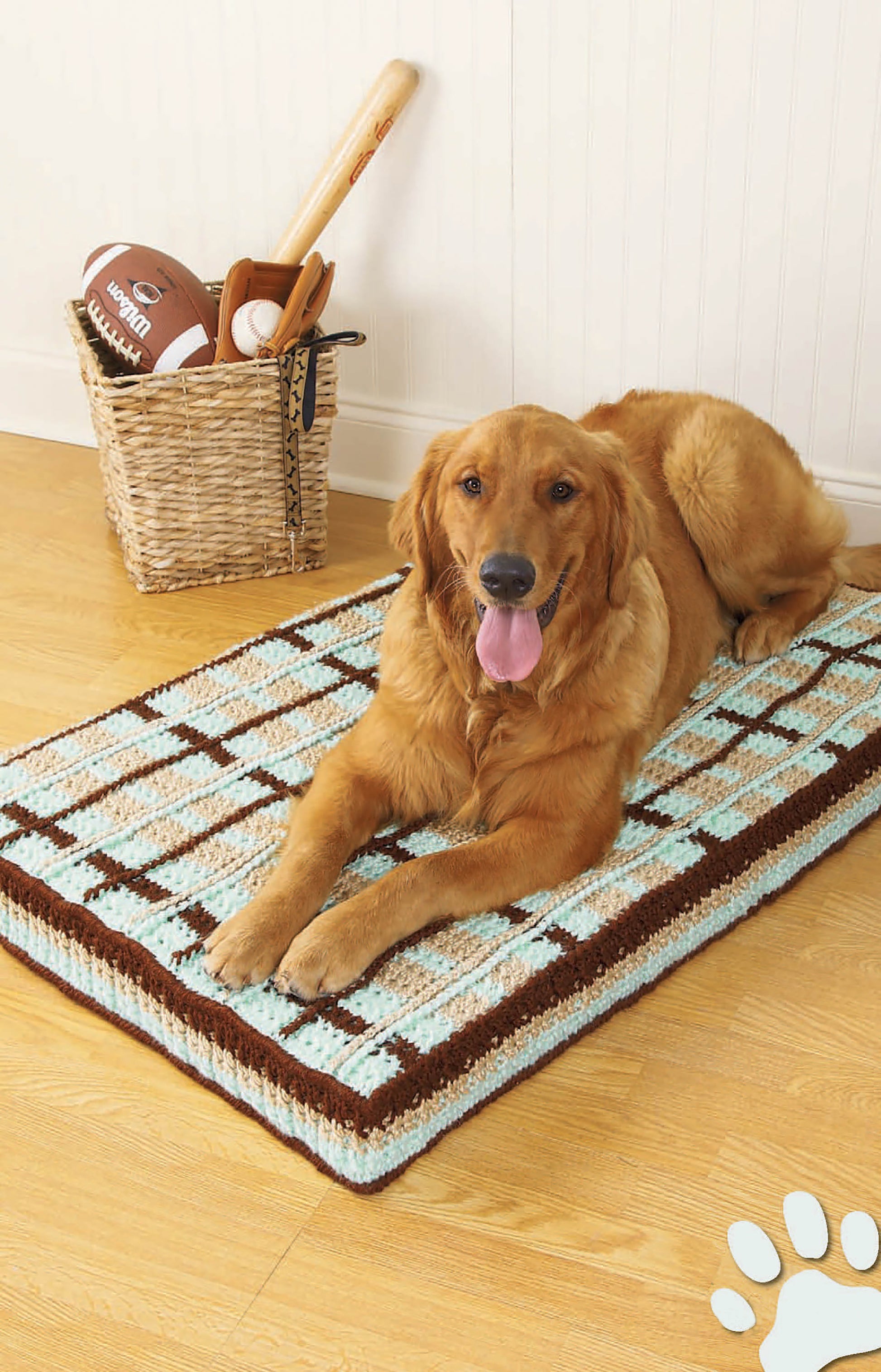 A golden retriever lounges by a cozy crochet bed crafted with medium weight yarn from the Sweet Pet Comforts Digital Download by Leisure Arts. Nearby, a basket holds sports gear; a white paw print design accents the image.