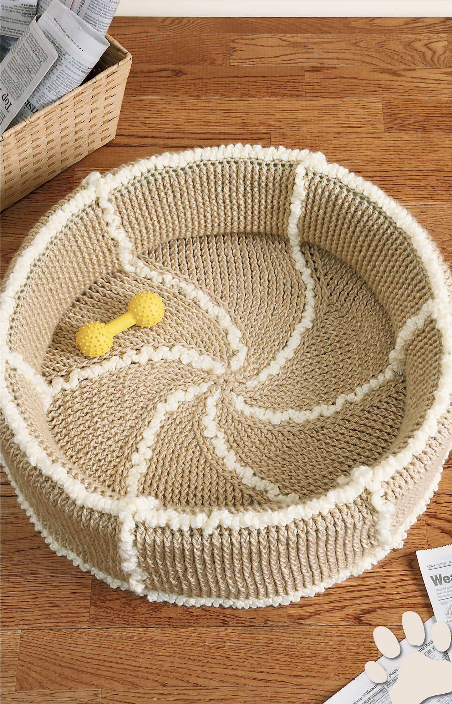 The Sweet Pet Comforts Digital Download by Leisure Arts features a round beige and white crochet pet bed on a wooden floor, with a yellow bone-shaped toy inside and a woven basket with newspapers nearby.