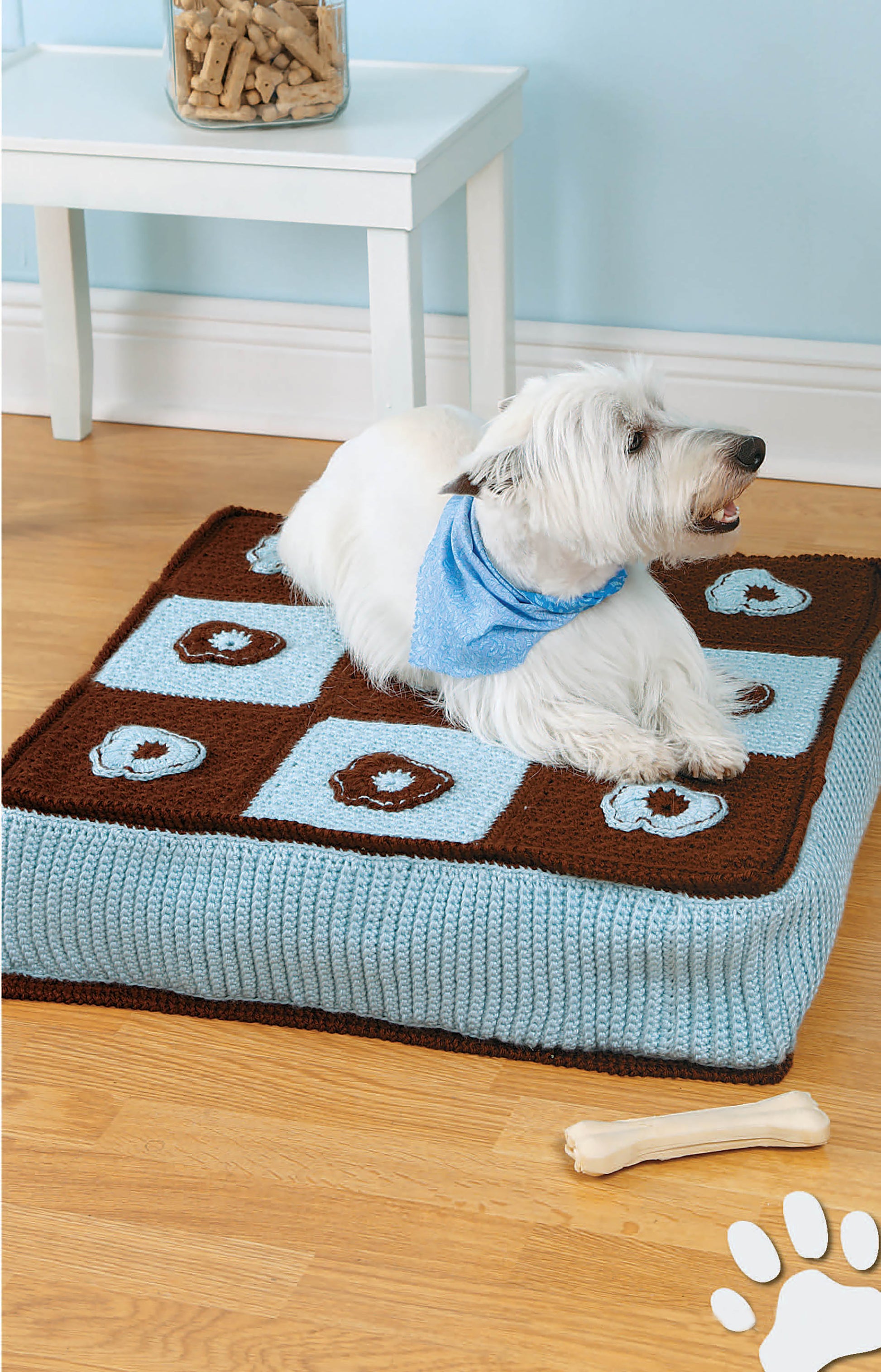 A small dog in a blue bandana rests on a blue and brown crochet bed made with medium weight yarn and a paw print design. Featured: Sweet Pet Comforts Digital Download by Leisure Arts. A jar of treats sits on a nearby table with a bone toy on the floor.