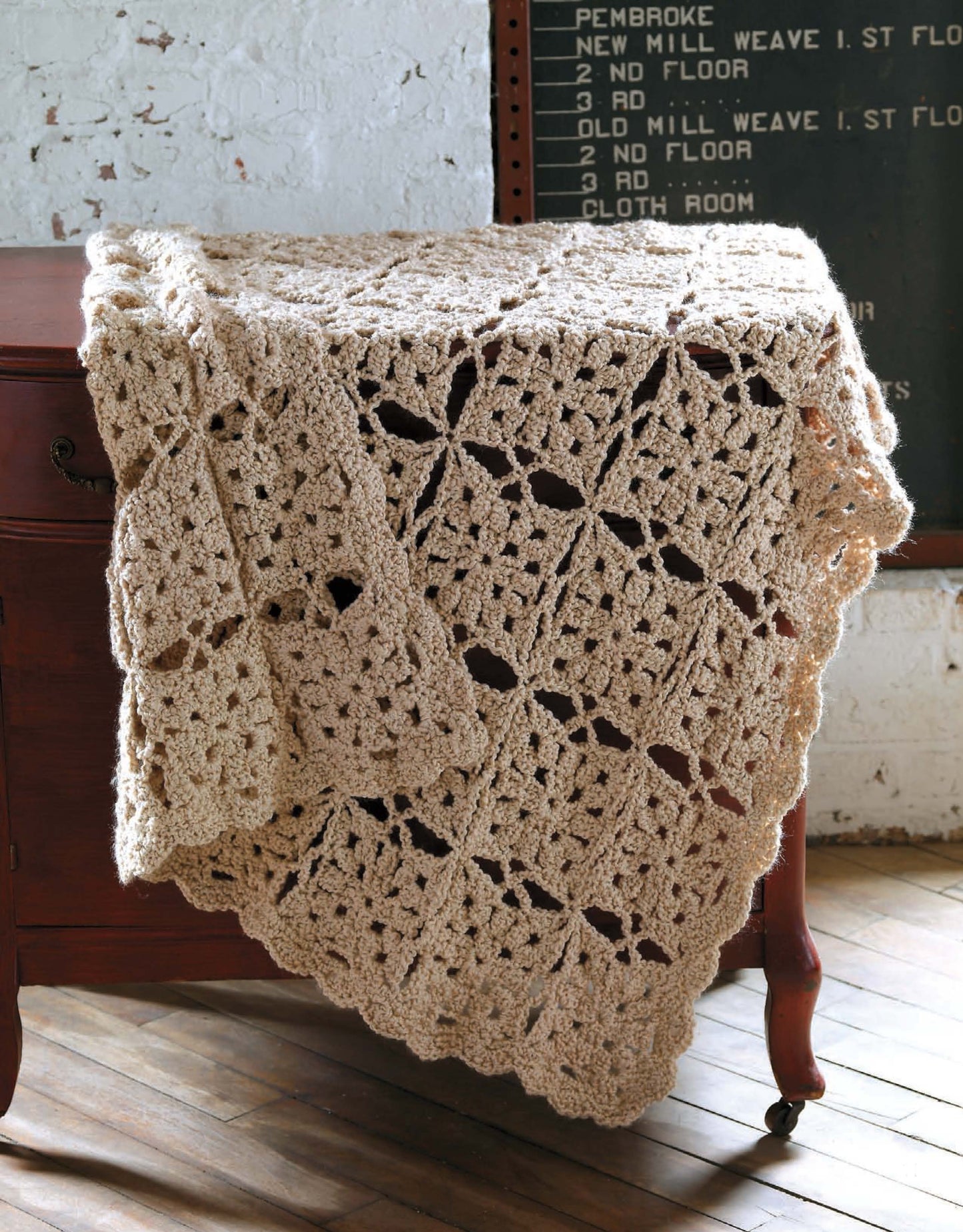 “The Story of Homespun Crochet” by Leisure Arts—a cream-toned, floral square afghan made with bulky yarn—rests on a wooden table, while a vintage sign decorates the white brick wall in the background.