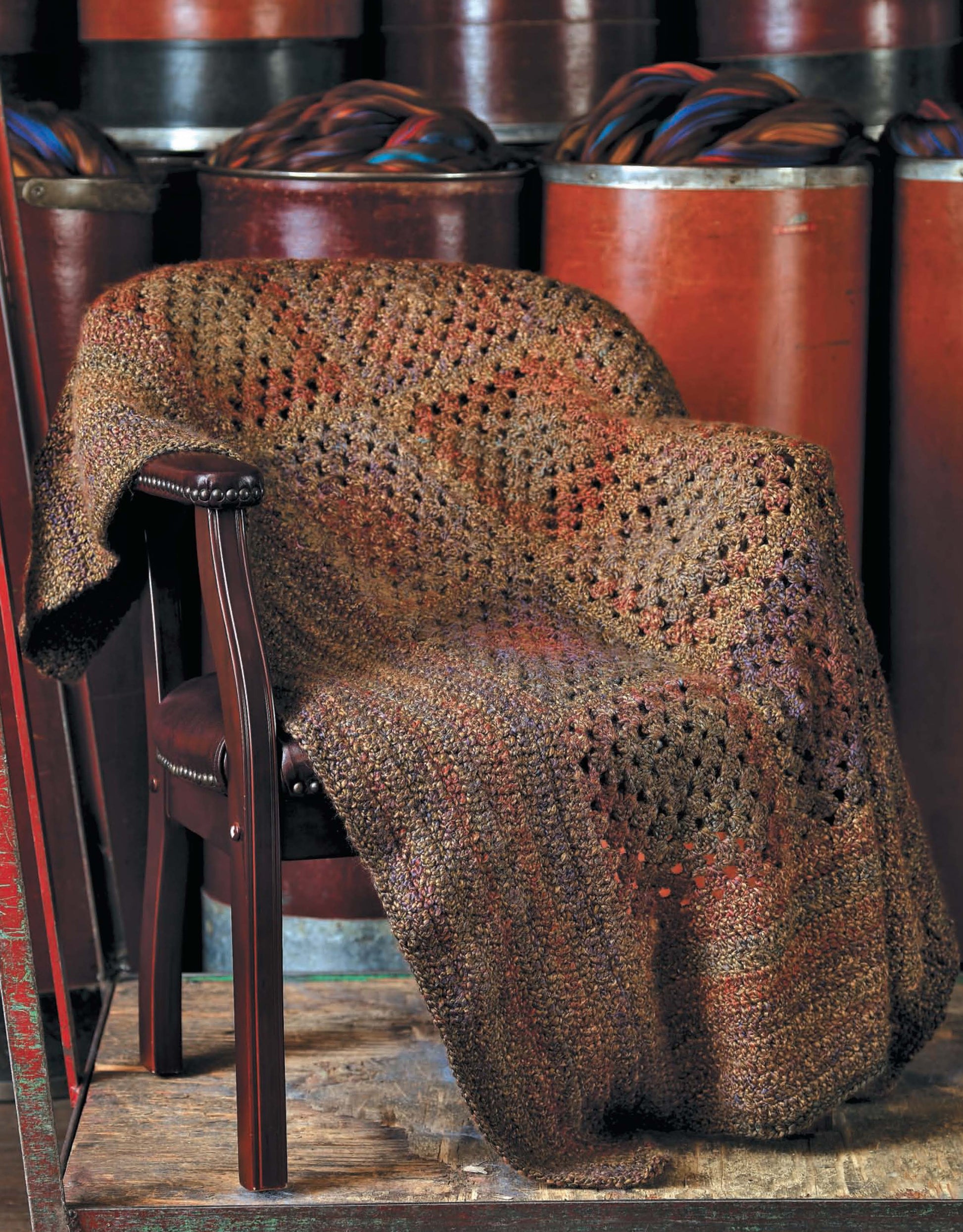 A brown upholstered chair displays an earth-toned crocheted blanket, made from bulky yarn using “The Story of Homespun Crochet” by Leisure Arts. In the background, tall red cylinders contain bundles of dark fiber.