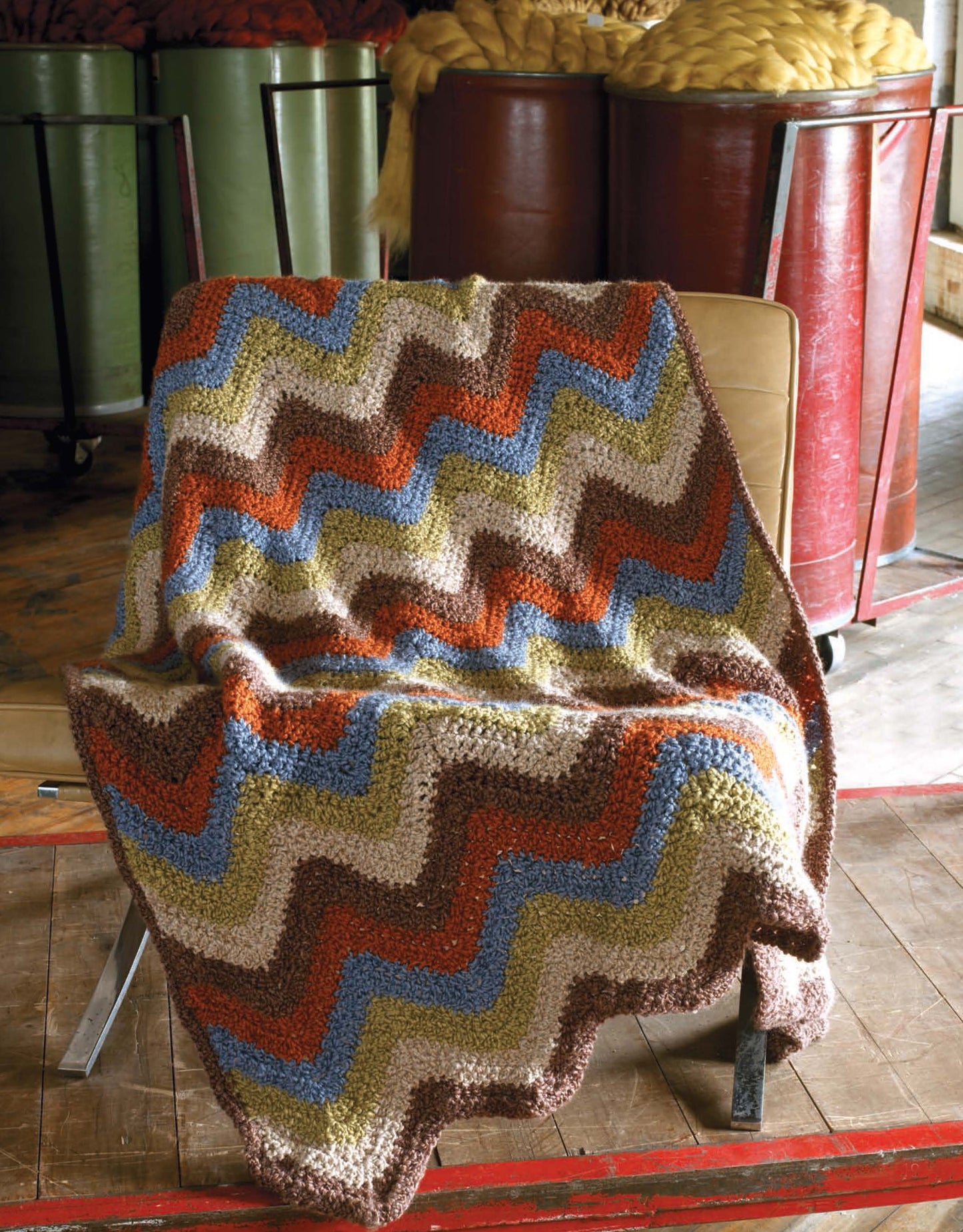 A vibrant zigzag-patterned blanket from Leisure Arts' "The Story of Homespun Crochet" rests on a tan chair, while large barrels of yellow wool sit in the background on a wooden workshop floor.