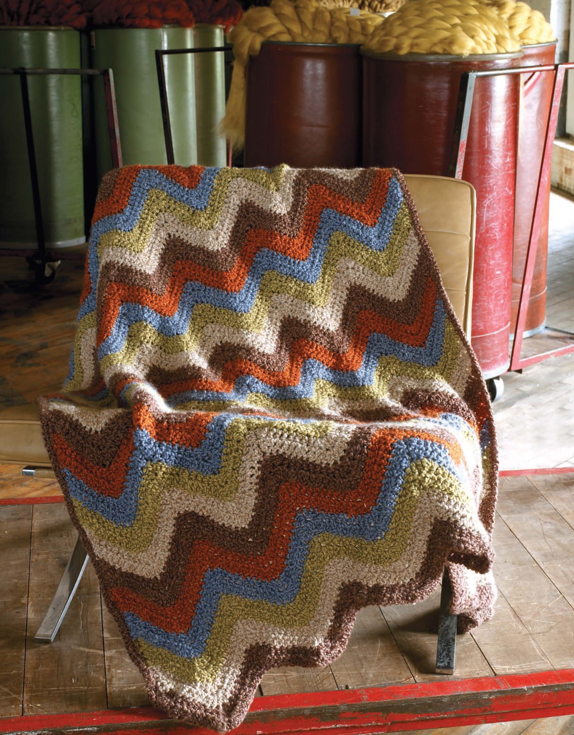 A vibrant zigzag-patterned blanket from Leisure Arts' "The Story of Homespun Crochet" rests on a tan chair, while large barrels of yellow wool sit in the background on a wooden workshop floor.