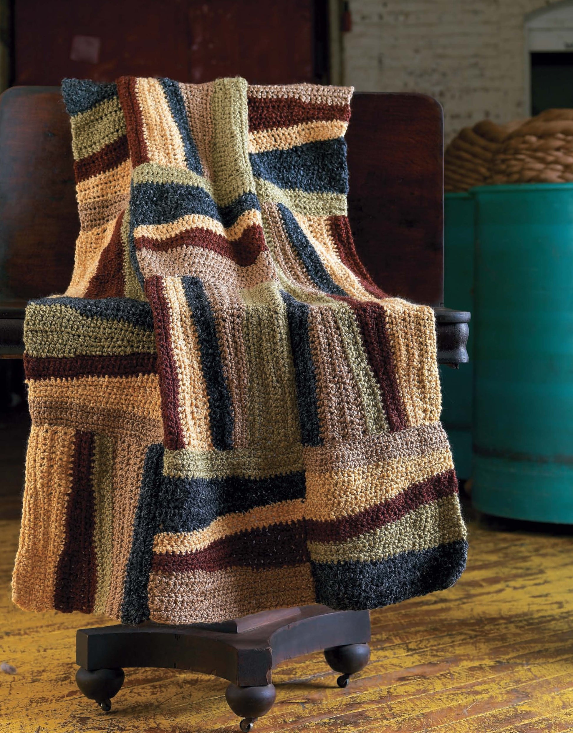 The Story of Homespun Crochet by Leisure Arts features a bulky-weight, knitted patchwork blanket draped over a dark wooden chair, set on a worn yellow floor with teal barrels and a brick wall in the background.