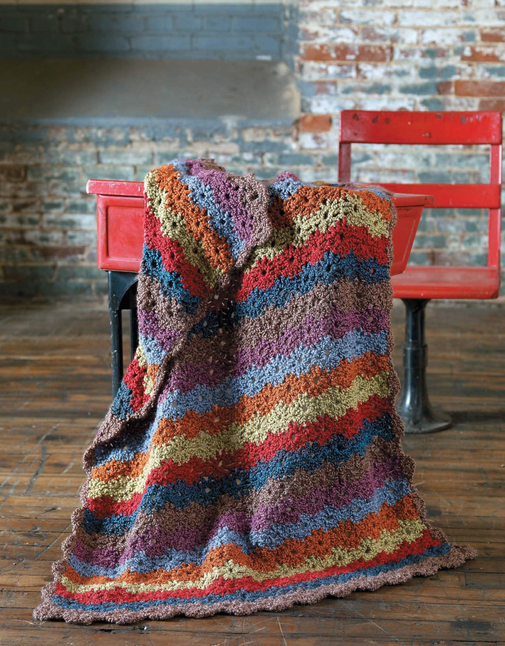 The Story of Homespun Crochet by Leisure Arts: A colorful, striped bulky yarn blanket is draped over a red wooden school desk and chair on a worn wooden floor with a rustic brick wall backdrop.