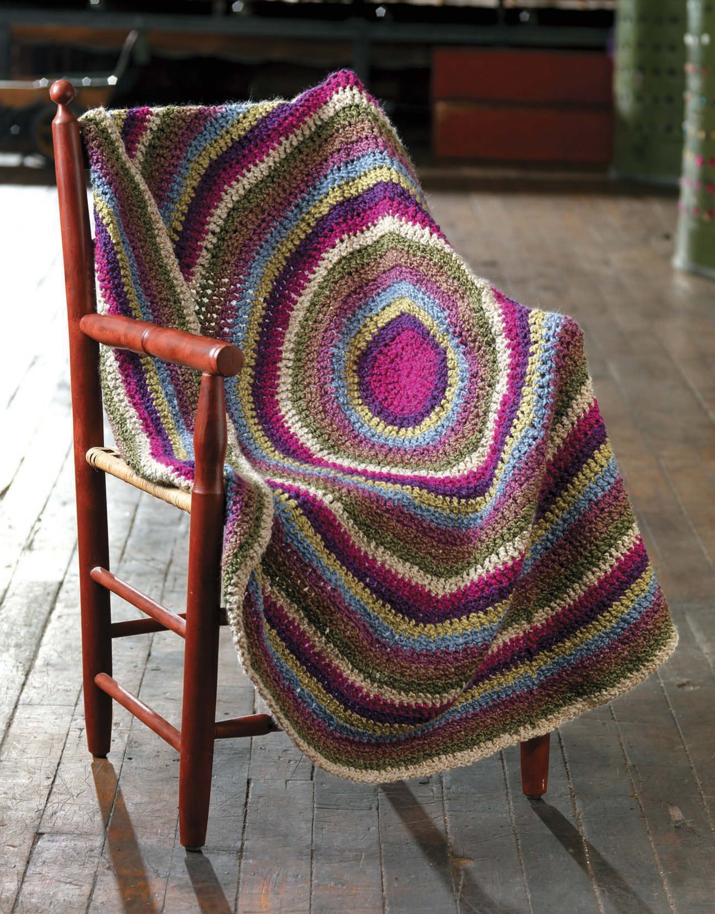 The Story of Homespun Crochet by Leisure Arts displays a cozy, colorful crocheted blanket with bulky yarn and concentric stripes in purple, pink, green, blue, and beige, draped over a wooden chair on a softly lit wood floor.