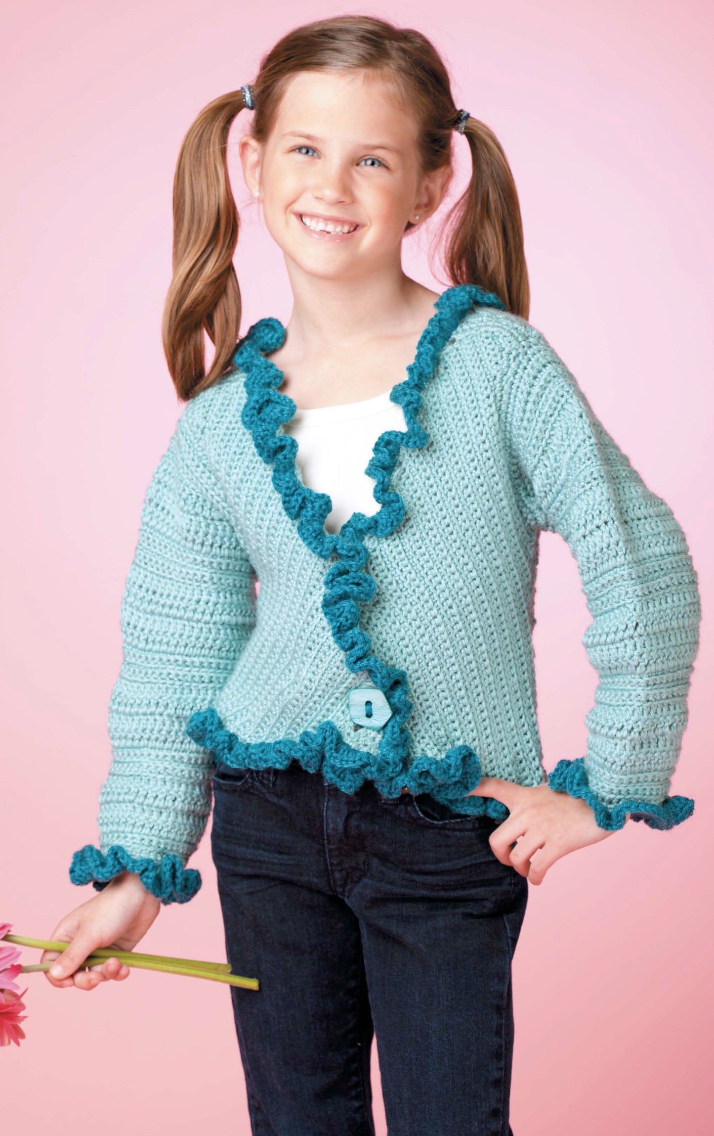 A smiling girl with brown pigtails models a light blue crocheted cardigan from Leisure Arts' Crochet Sweaters & Dresses for Girls collection, standing against a pink backdrop and holding pink flowers.