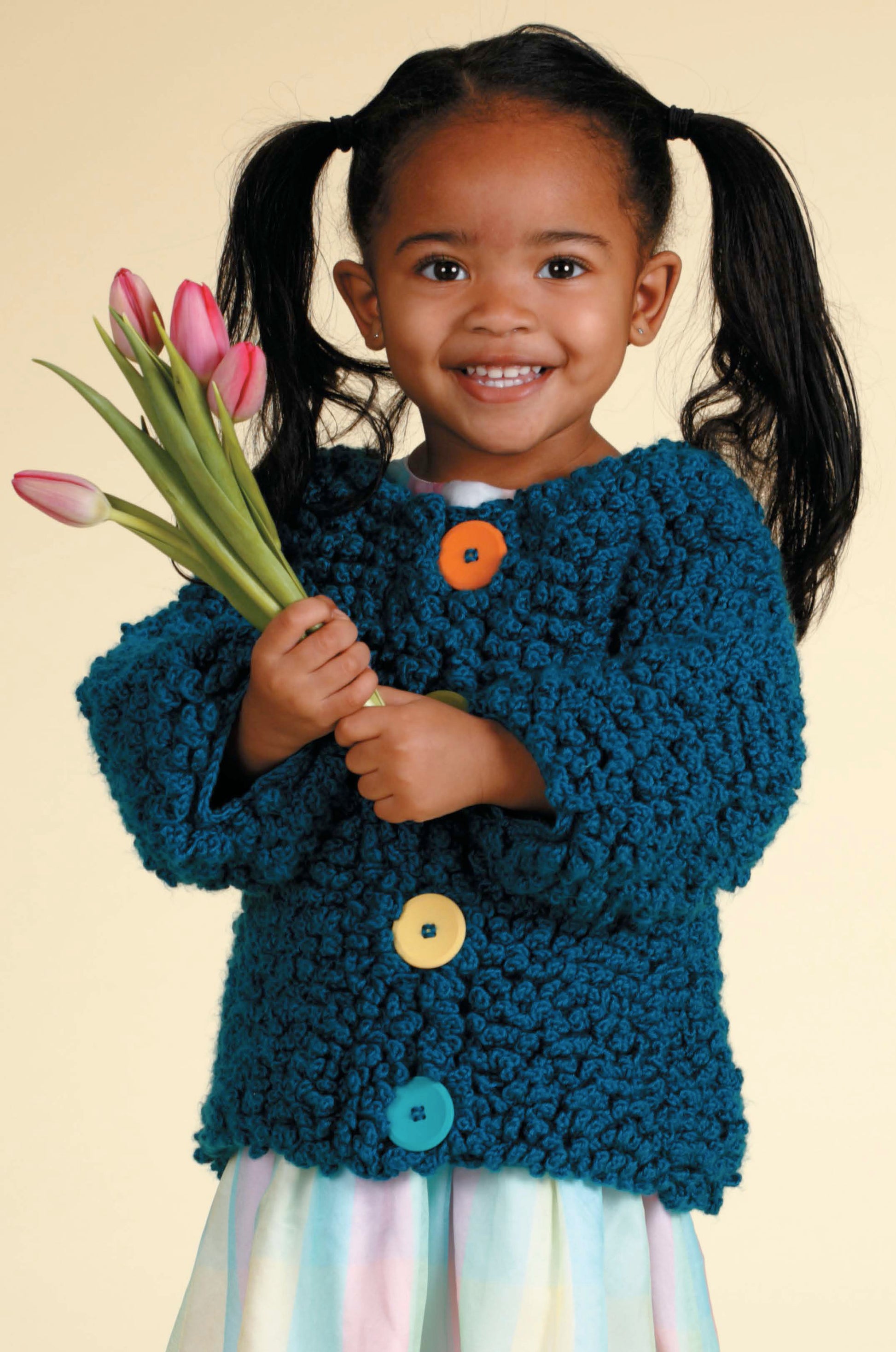 A smiling girl with pigtails holds pink tulips, wearing a teal cardigan from Leisure Arts' "Crochet Sweaters & Dresses for Girls"—a perfect inspiration for handcrafted children’s fashion. She pairs it with a pastel striped skirt.