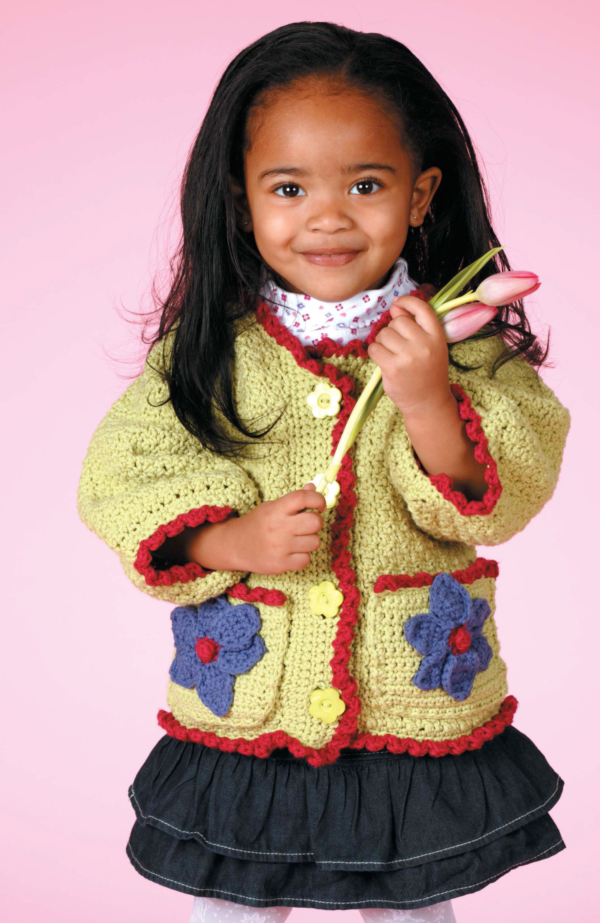 A young girl in Leisure Arts’ Crochet Sweaters & Dresses for Girls—a yellow floral sweater and black skirt—smiles holding two pink tulips against a pink background.