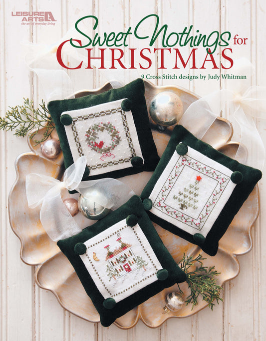 Three green velvet Christmas ornaments featuring cross stitch designs—a wreath, tree, and house—are displayed on a white rustic surface. Text reads Sweet Nothings Cross Stitch for Christmas by Leisure Arts.