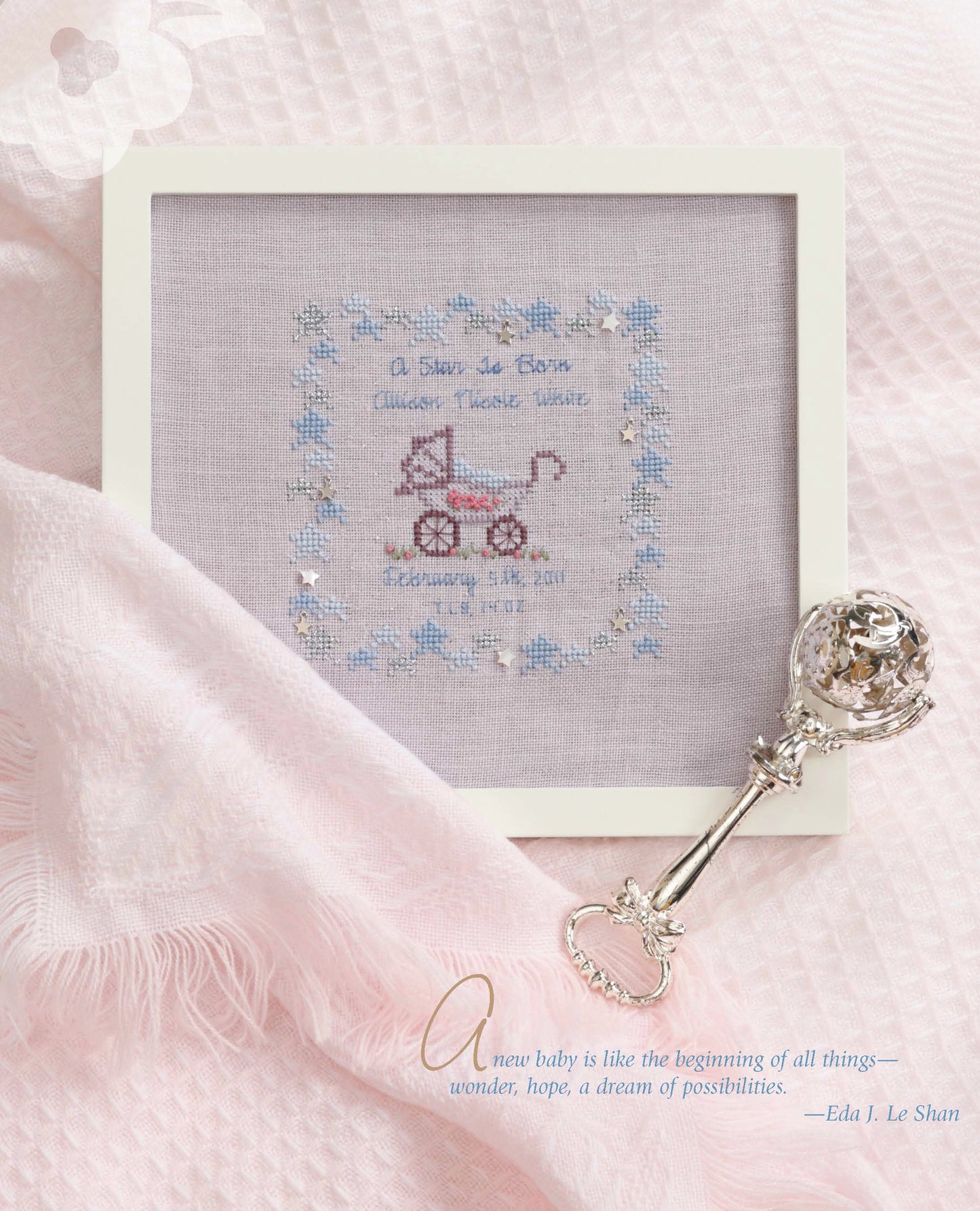 The Sweet Nothings Cross Stitch for Baby by Leisure Arts is a framed project with a pink rocking horse and blue floral border, displayed on a pink blanket next to a silver rattle. A quote about new babies and possibilities appears at the bottom right.