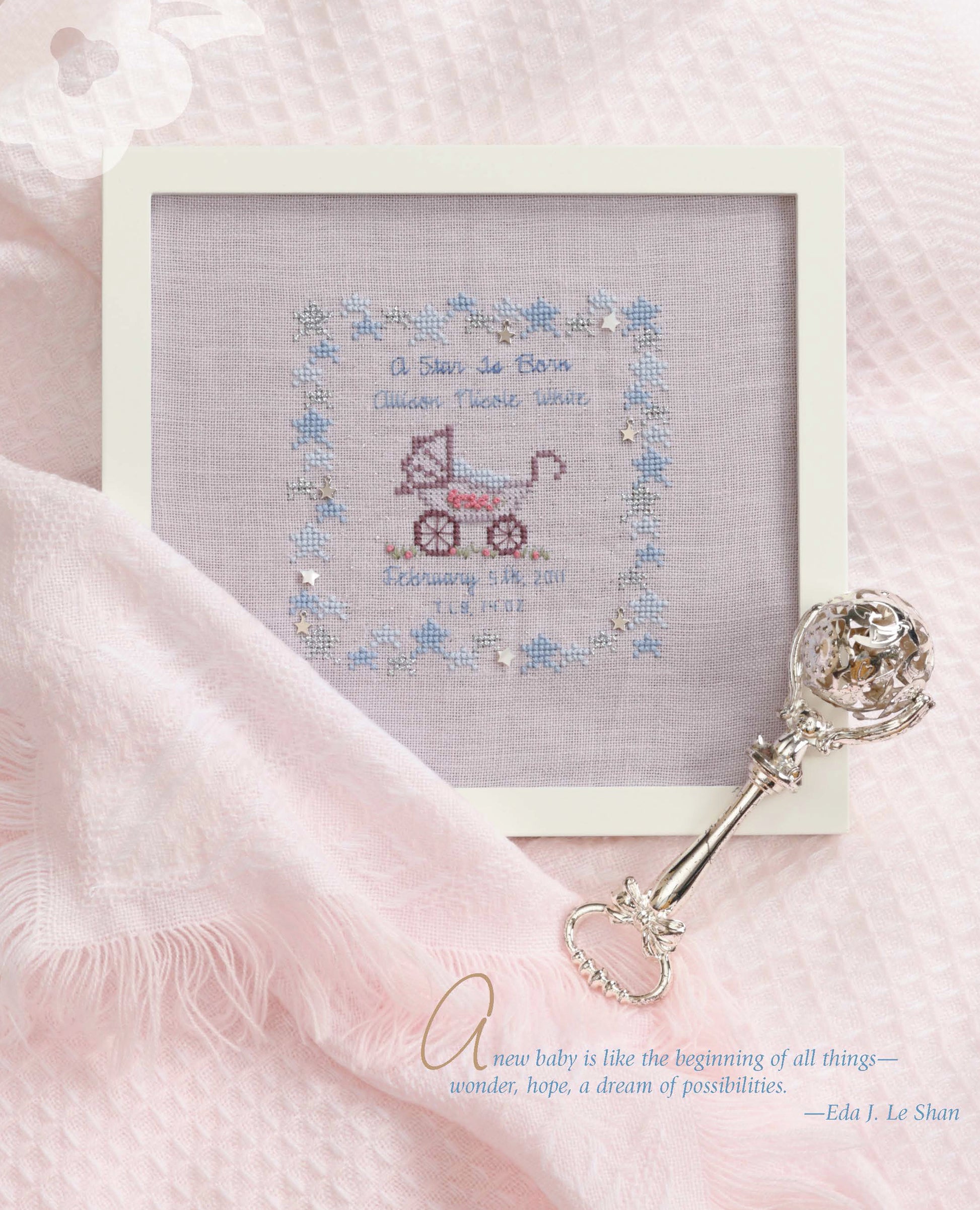 The Sweet Nothings Cross Stitch for Baby by Leisure Arts is a framed project with a pink rocking horse and blue floral border, displayed on a pink blanket next to a silver rattle. A quote about new babies and possibilities appears at the bottom right.