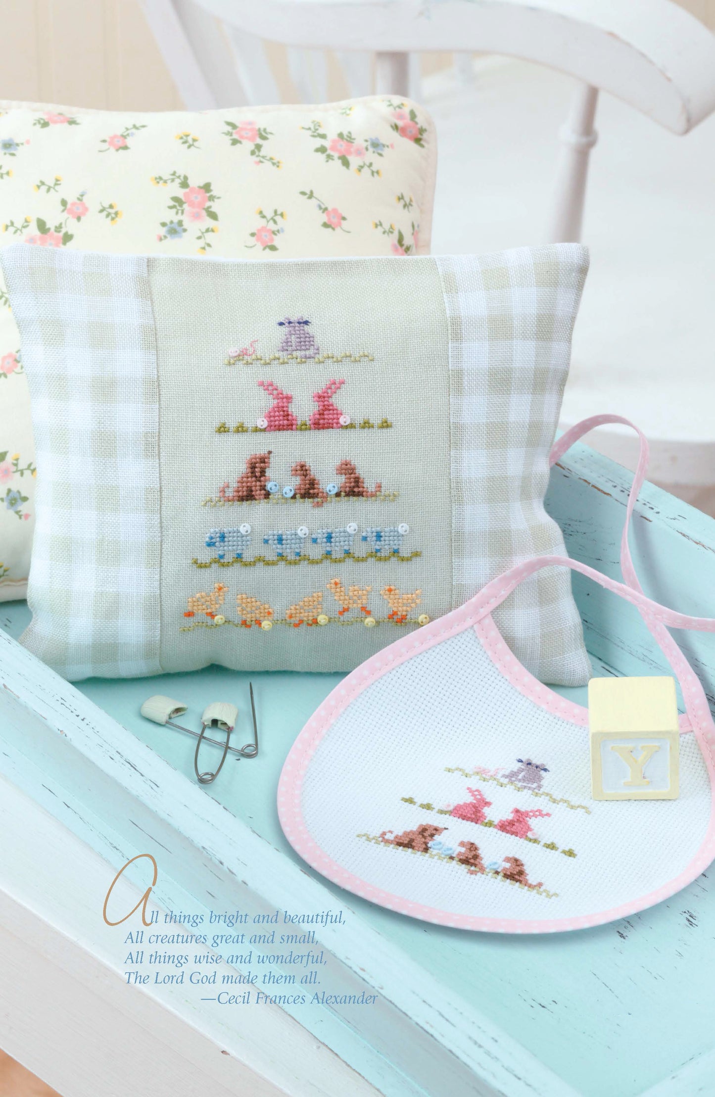Displayed on a pale blue tray are items made with Leisure Arts' Sweet Nothings Cross Stitch for Baby, including a small pillow and baby bib with pastel animal designs, plus safety pins and a yellow pacifier; a floral cushion rests in the background.