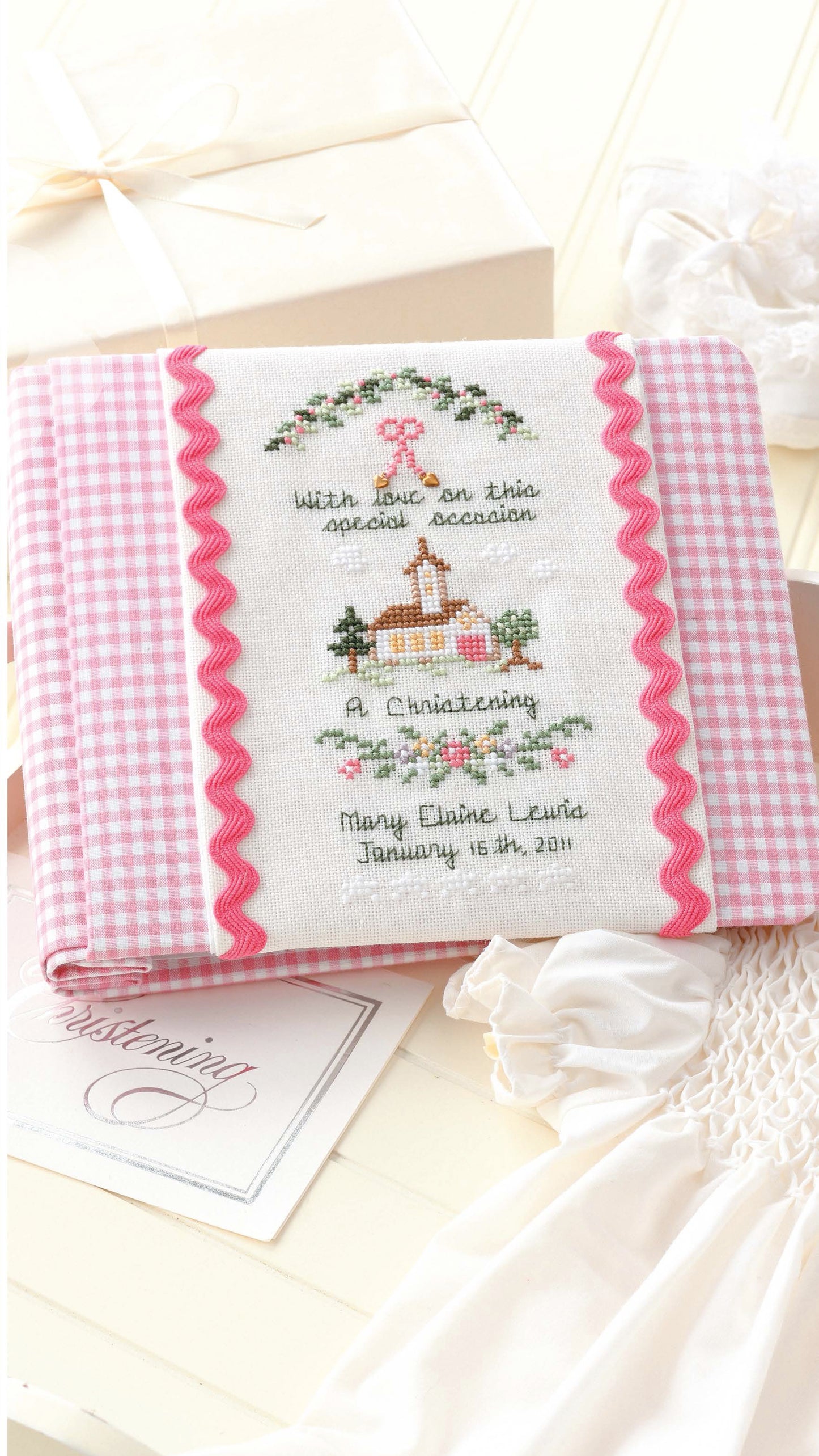 The "Sweet Nothings Cross Stitch for Baby" by Leisure Arts features a pink gingham photo album with an embroidered church and floral motifs, personalized for christening—a lovely cross stitch inspiration among cards, a white dress, and a gift box.
