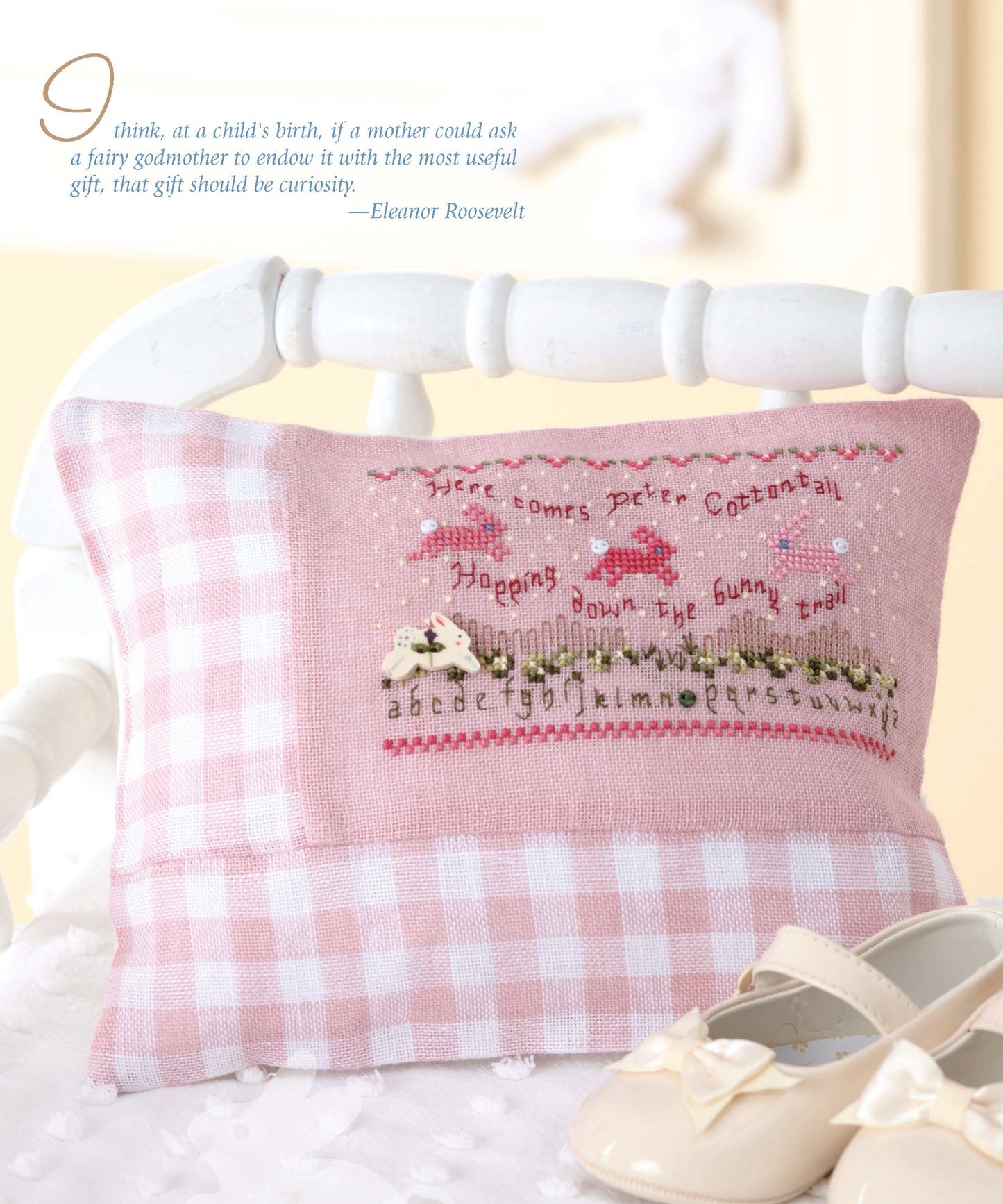 The “Sweet Nothings Cross Stitch for Baby” by Leisure Arts features a pink gingham pillow with an embroidered bunny scene and alphabet, displayed beside white baby shoes and a curiosity quote by Eleanor Roosevelt in the upper left corner.