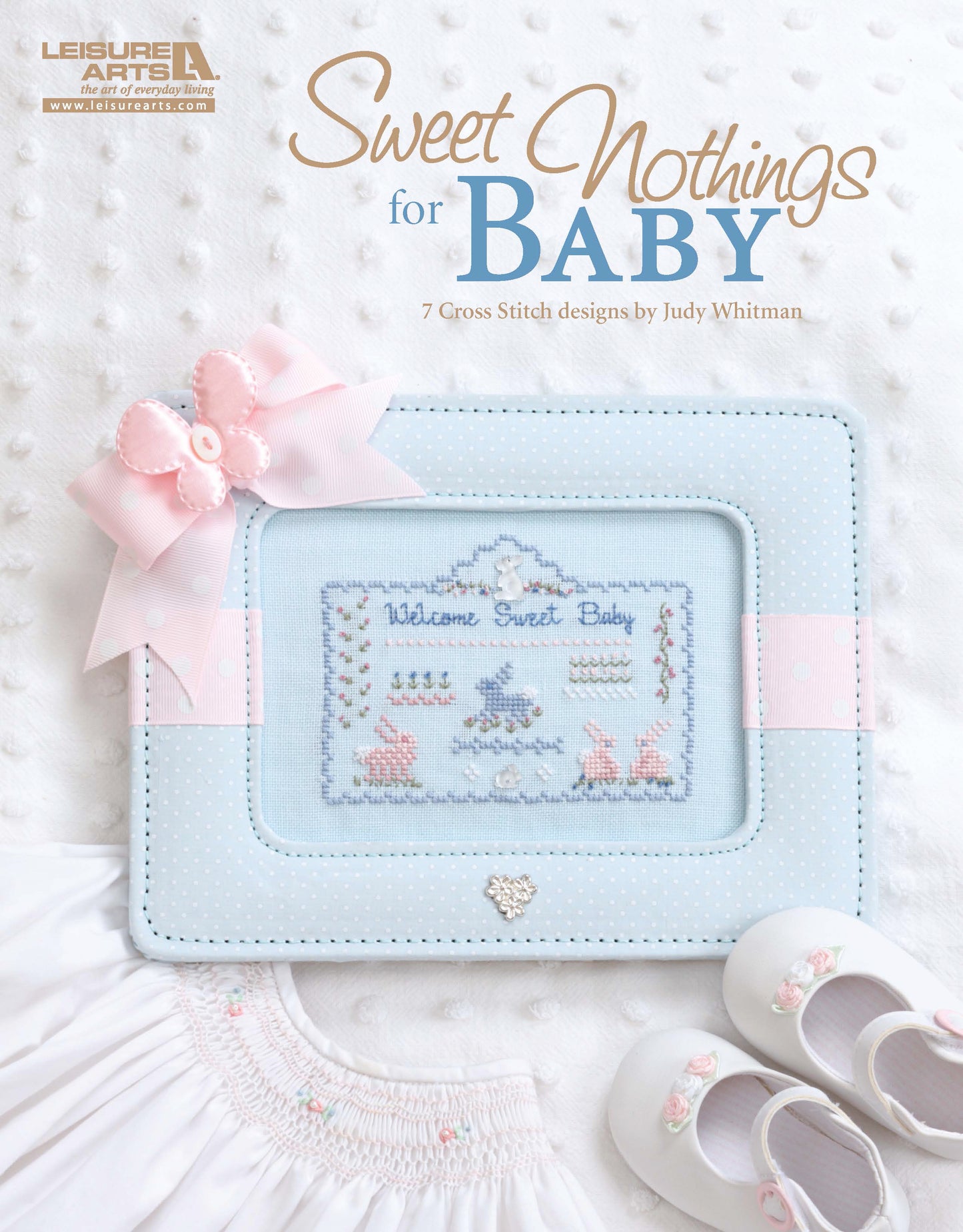 A pastel blue frame with counted cross stitch from Leisure Arts’ Sweet Nothings Cross Stitch for Baby features welcome baby text and pink and blue motifs, displayed beside baby shoes with pink bows. Text: Sweet Nothings for Baby.