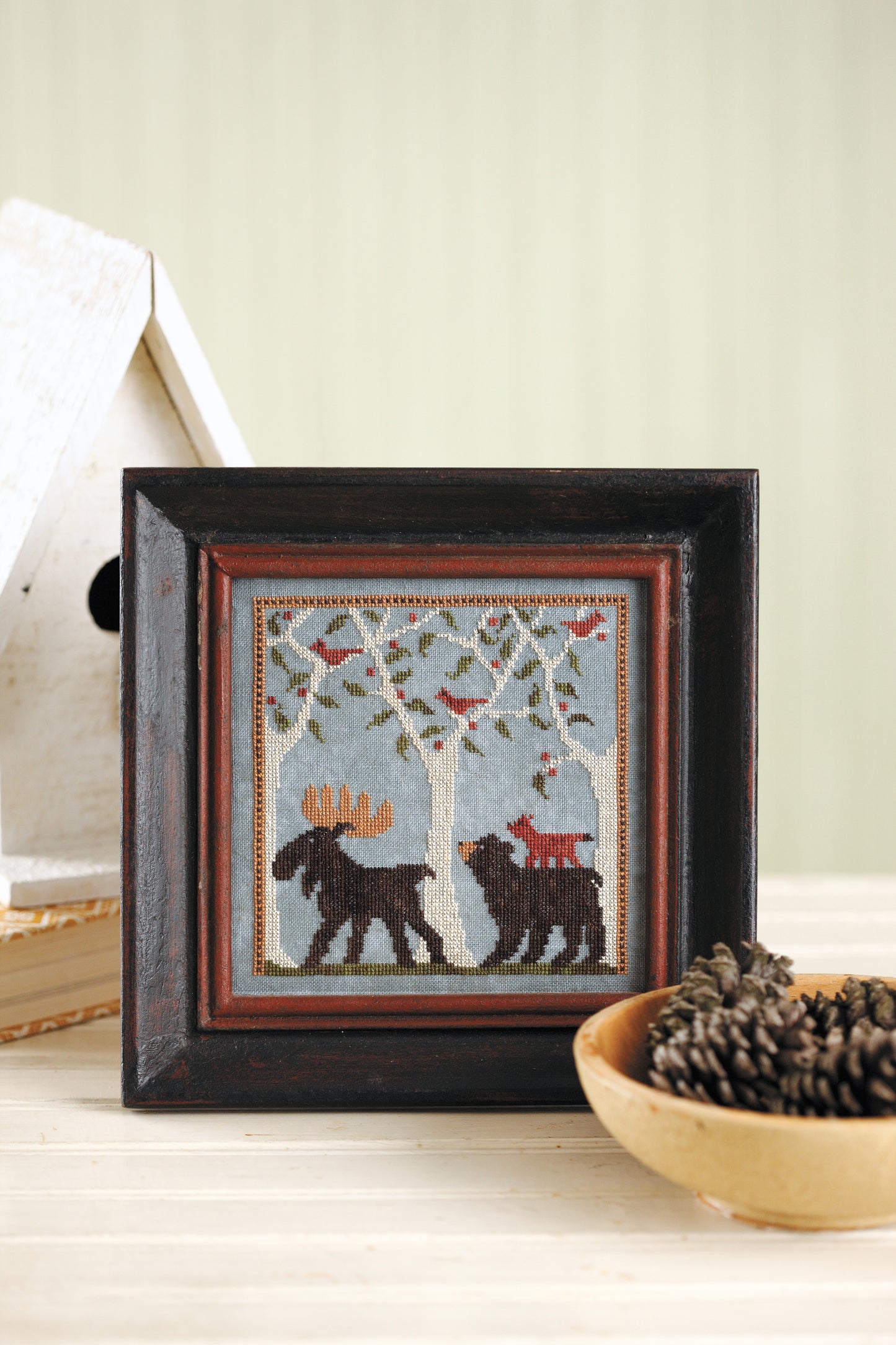 The Leisure Arts "All Creatures Stitched & Small" cross stitch by Lori Markovic features two black bears under leafy branches with red and orange birds perched above. It's displayed on a light table beside a white birdhouse and a wooden bowl of pinecones.