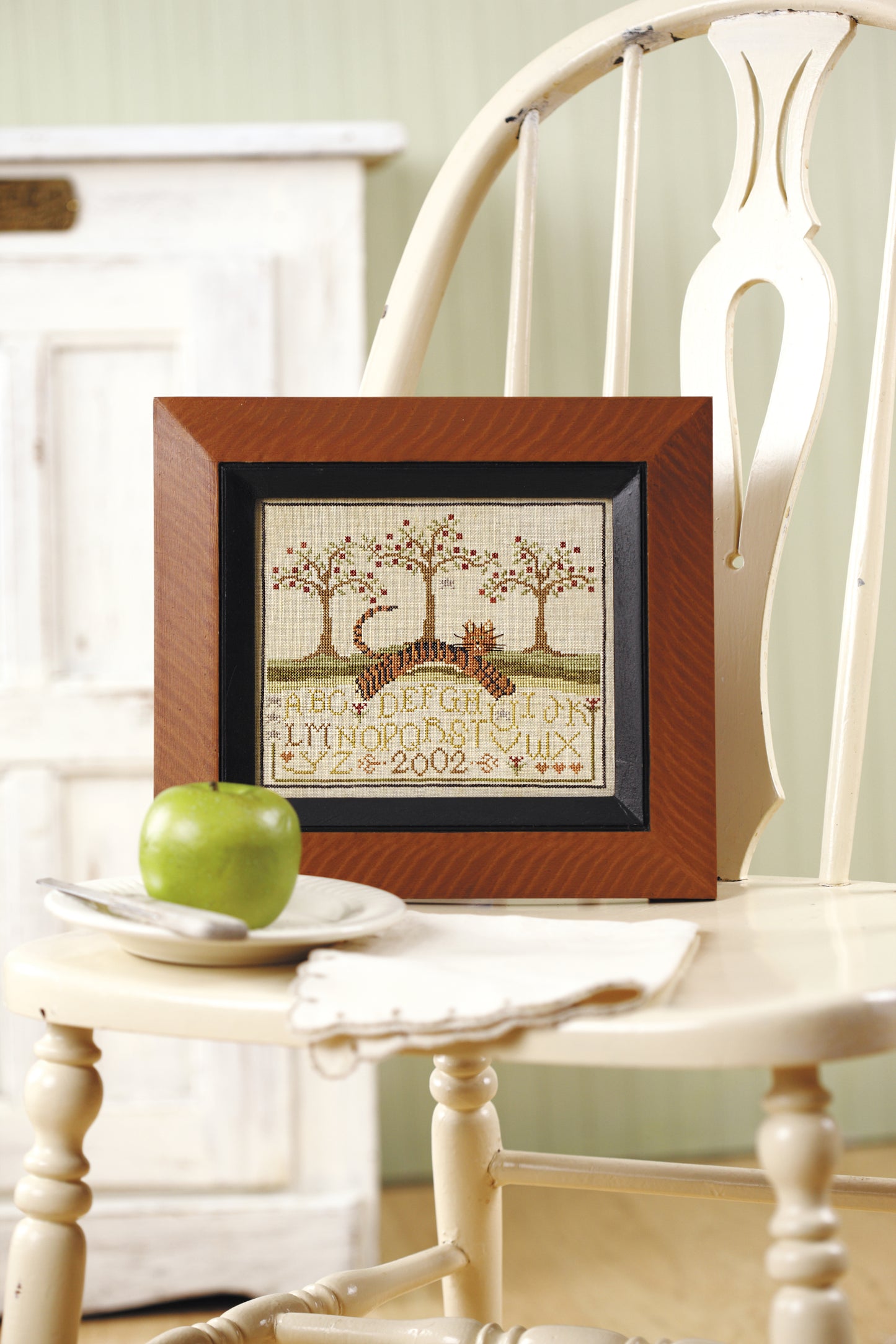 The “All Creatures Stitched & Small” counted cross stitch sampler by Leisure Arts, featuring trees, a deer, alphabet letters, and “2002,” is framed and displayed on a white chair beside a green apple and white plate; a cabinet is behind.