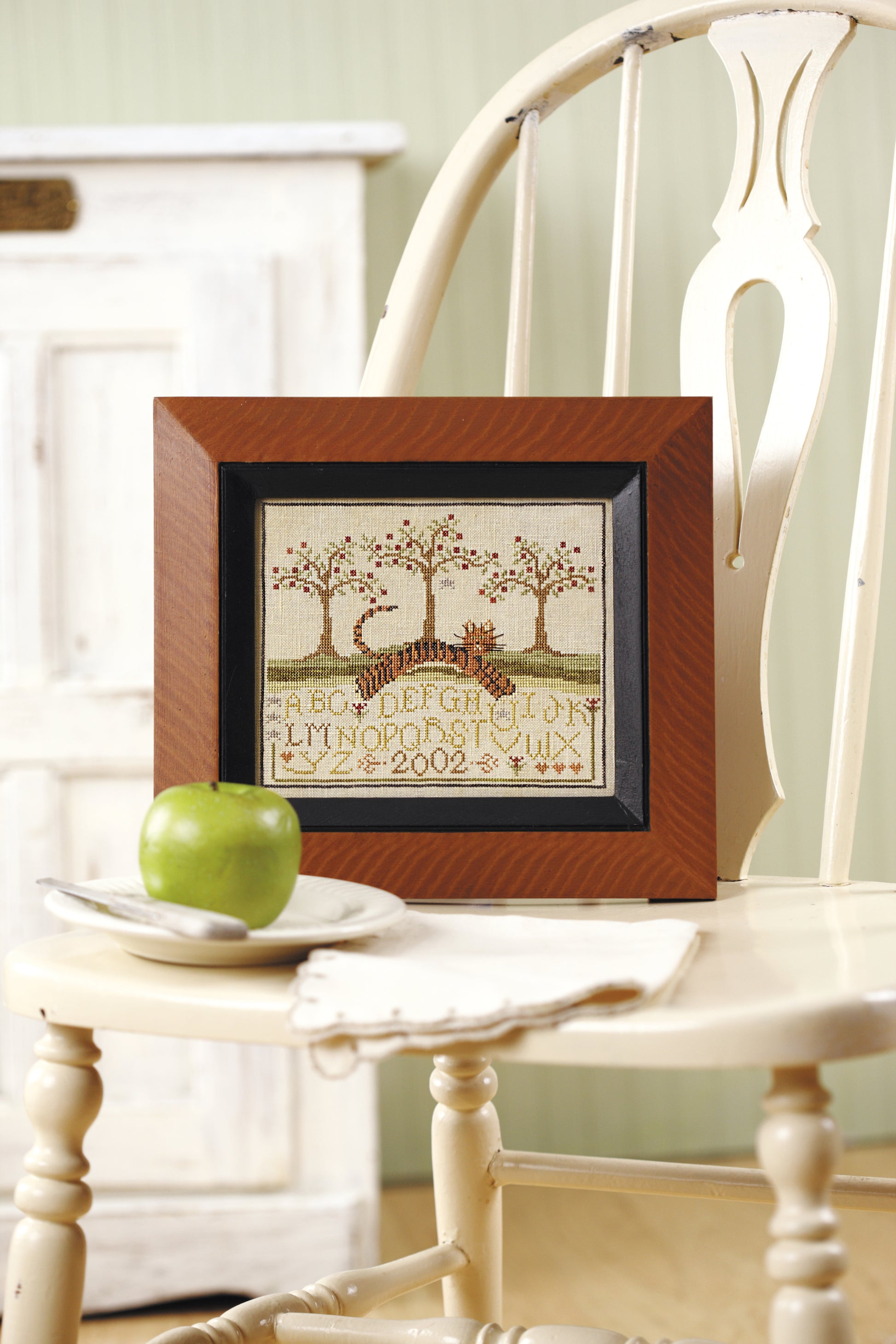 The “All Creatures Stitched & Small” counted cross stitch sampler by Leisure Arts, featuring trees, a deer, alphabet letters, and “2002,” is framed and displayed on a white chair beside a green apple and white plate; a cabinet is behind.