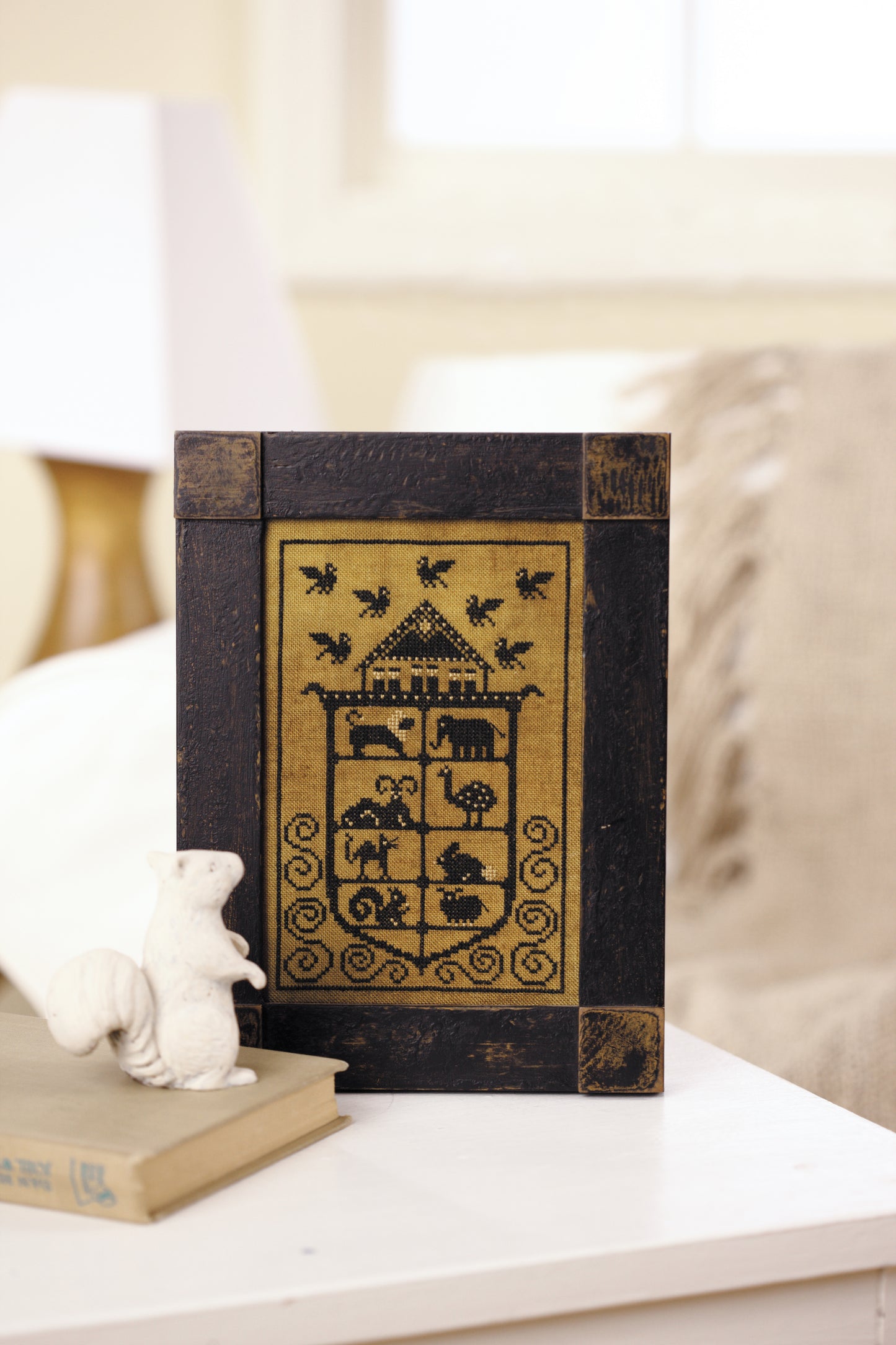 The framed “All Creatures Stitched & Small” counted cross stitch by Leisure Arts rests on a white table beside a ceramic squirrel and closed book, with animals, a house, and birds depicted against a softly lit, cozy background.