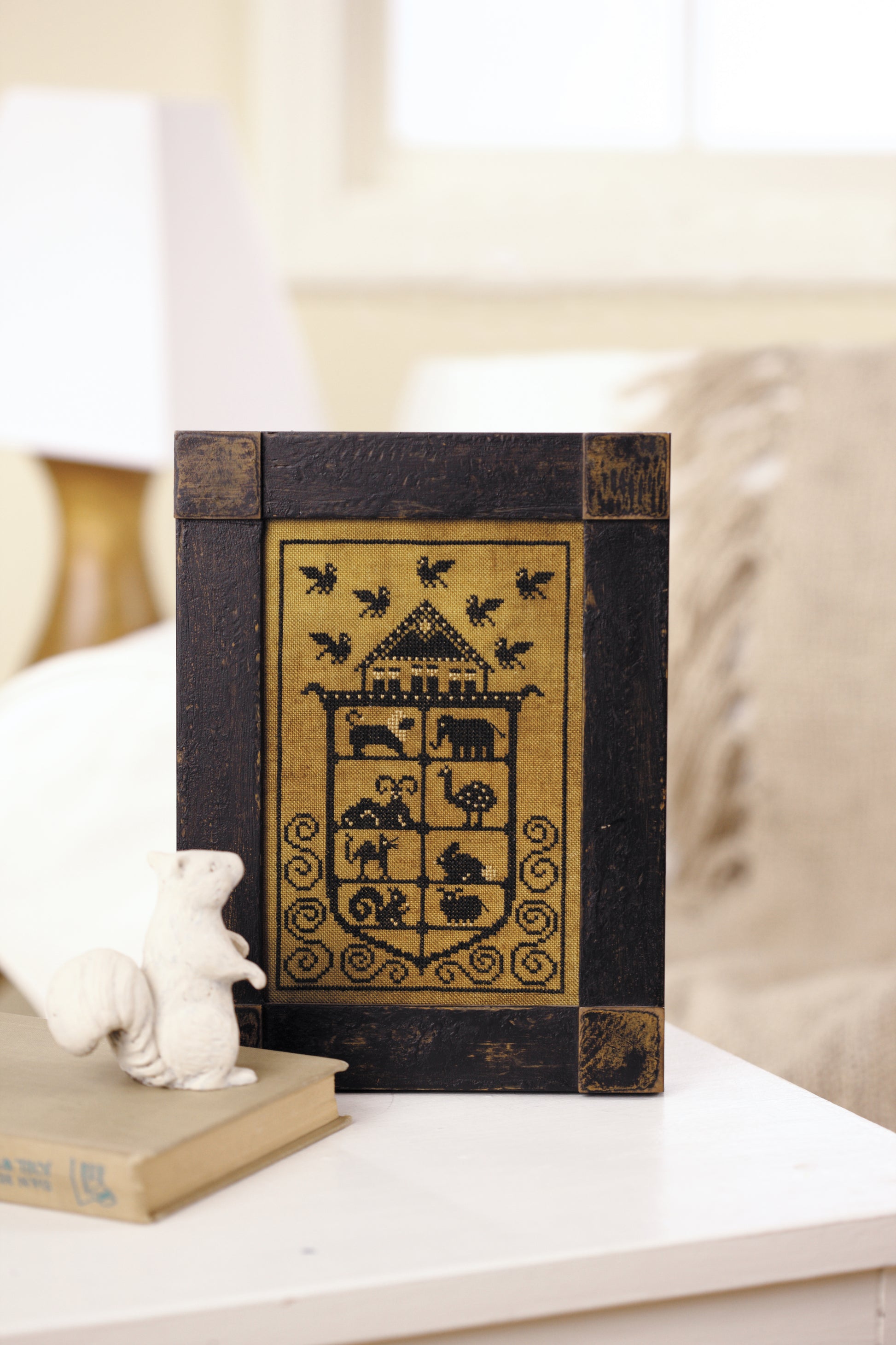 The framed “All Creatures Stitched & Small” counted cross stitch by Leisure Arts rests on a white table beside a ceramic squirrel and closed book, with animals, a house, and birds depicted against a softly lit, cozy background.