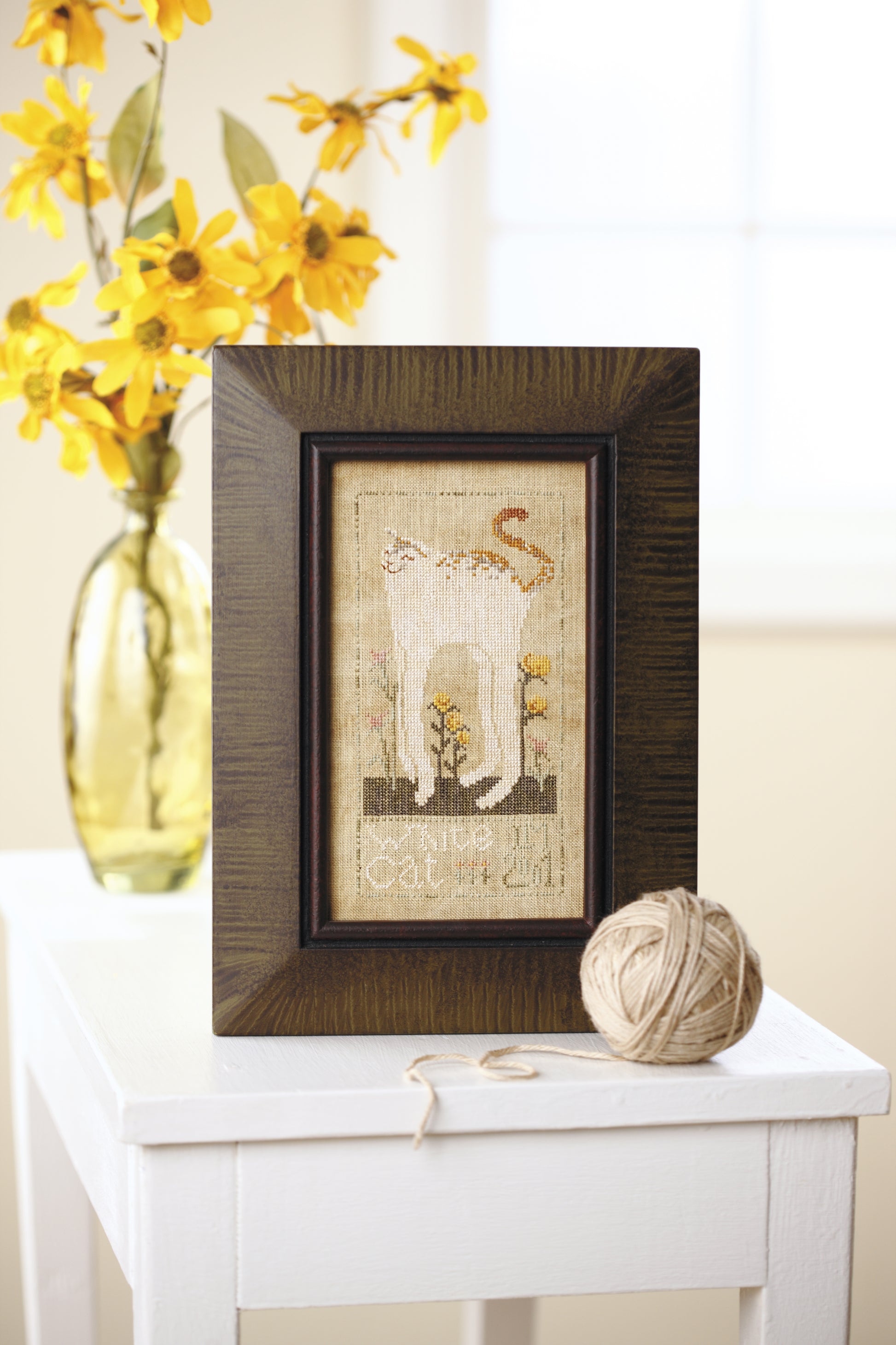 A framed counted cross stitch from Leisure Arts' "All Creatures Stitched & Small" shows a white cat with its tail raised on a white table near twine, with yellow flowers in a glass vase glowing by a sunlit window.