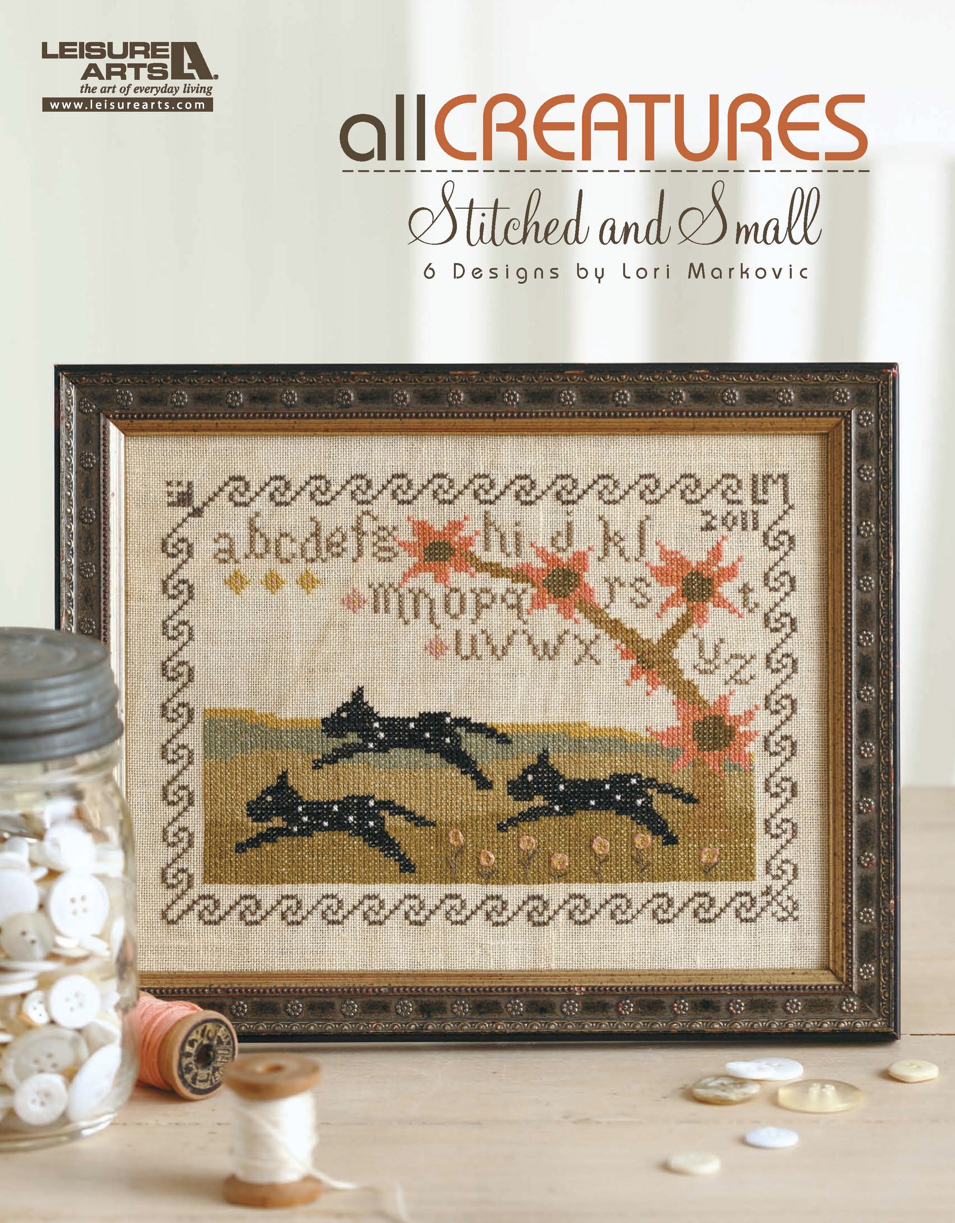 All Creatures Stitched & Small by Leisure Arts is framed counted cross stitch art showing two black dogs under an autumn tree, bordered by alphabet and patterns, with buttons and thread spools in front and title text above.