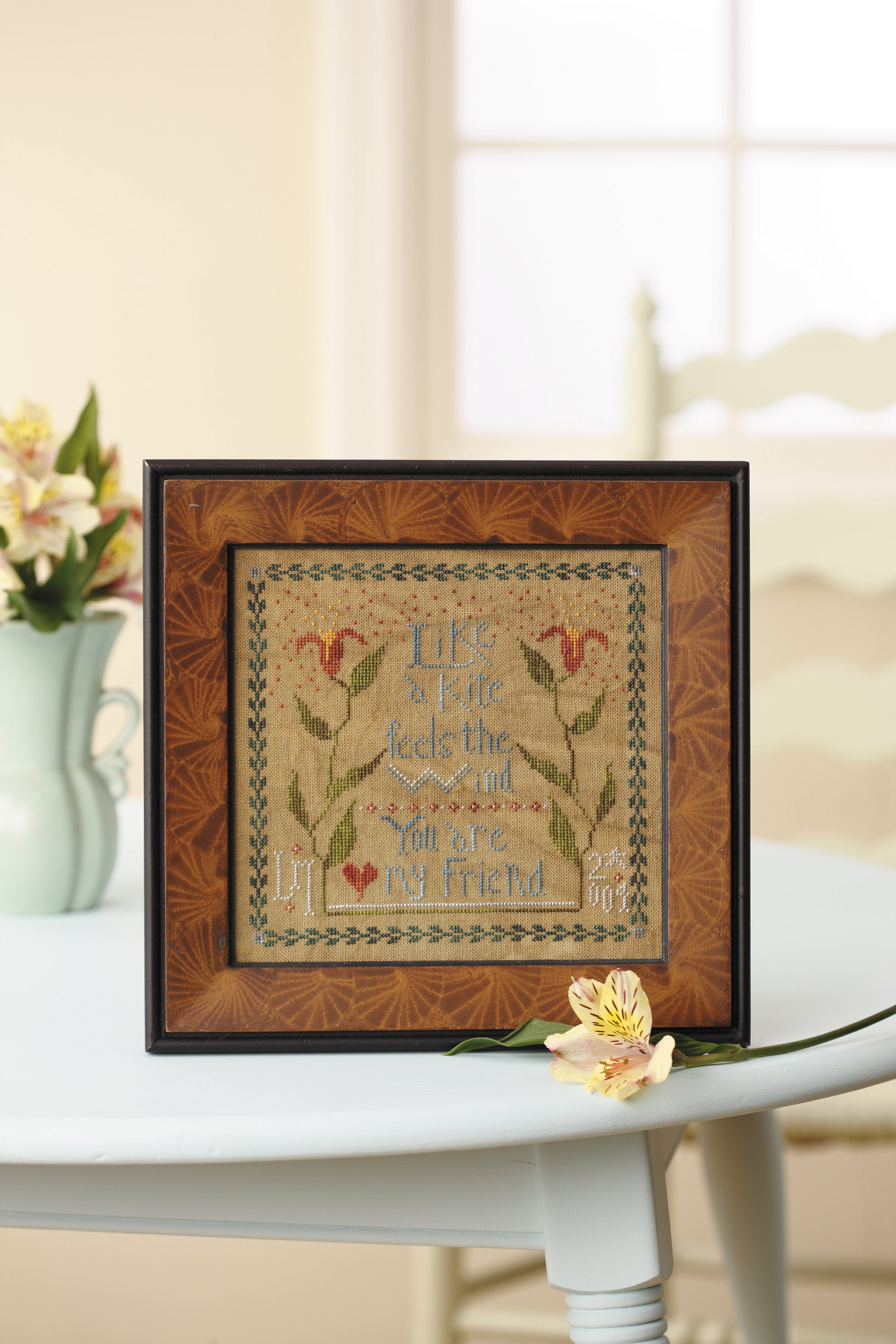 The "Words To Stitch By" cross-stitch by Leisure Arts, featuring the phrase “Life is a gift, tied with love, and you are my friend,” is displayed on a round white table near a yellow flower and a vase of flowers.