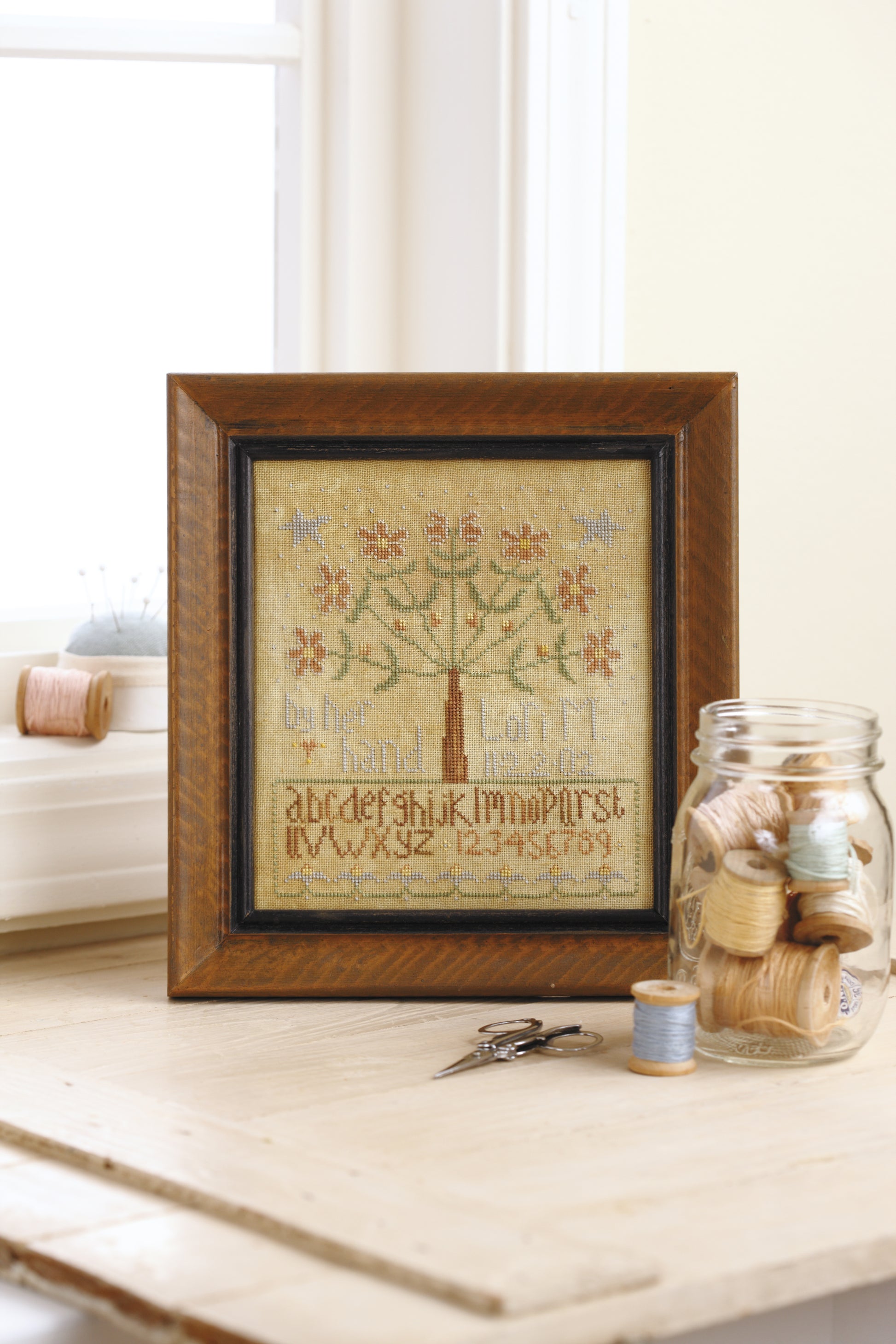 The "Words To Stitch By" cross-stitch sampler by Leisure Arts features an alphabet, numbers, and floral tree, displayed on a table with sewing tools—ideal for anyone who loves beautiful needlework and creative patterns.