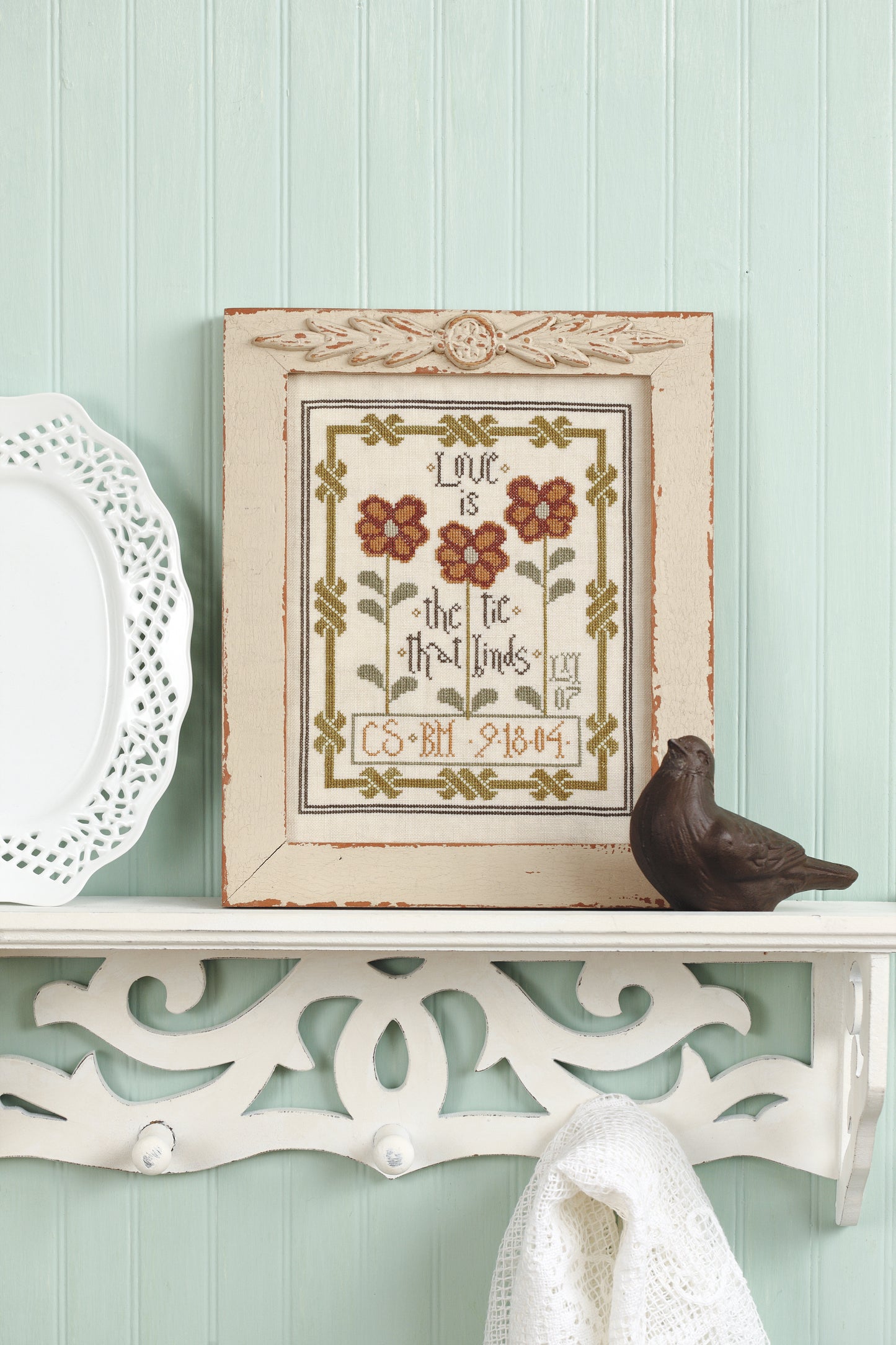 The Leisure Arts "Words To Stitch By" cross-stitch, with red flowers and “Love is the tie that binds,” designed by Lori Markovic, is displayed on a white shelf beside a ceramic bird and plate against a light green paneled wall.