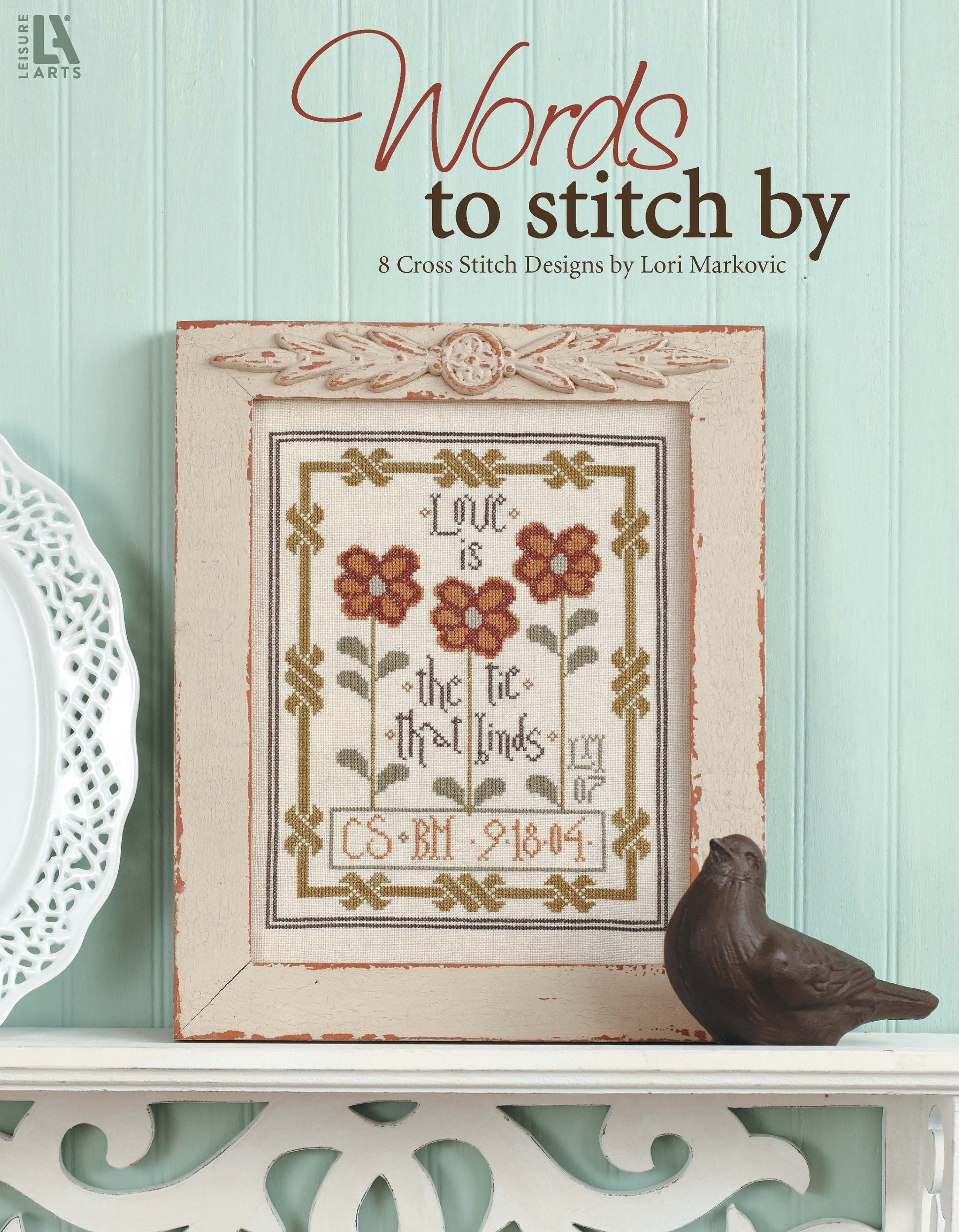 A framed floral cross-stitch from Leisure Arts’ “Words To Stitch By” sits on a white shelf against a mint-green wall, accompanied by a brown bird figurine. Text above states: “Words To Stitch By, 8 Cross Stitch Designs by Lori Markovic.”.