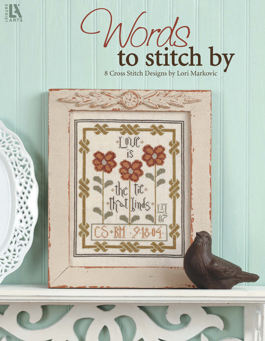 A framed floral cross-stitch from Leisure Arts’ “Words To Stitch By” sits on a white shelf against a mint-green wall, accompanied by a brown bird figurine. Text above states: “Words To Stitch By, 8 Cross Stitch Designs by Lori Markovic.”.