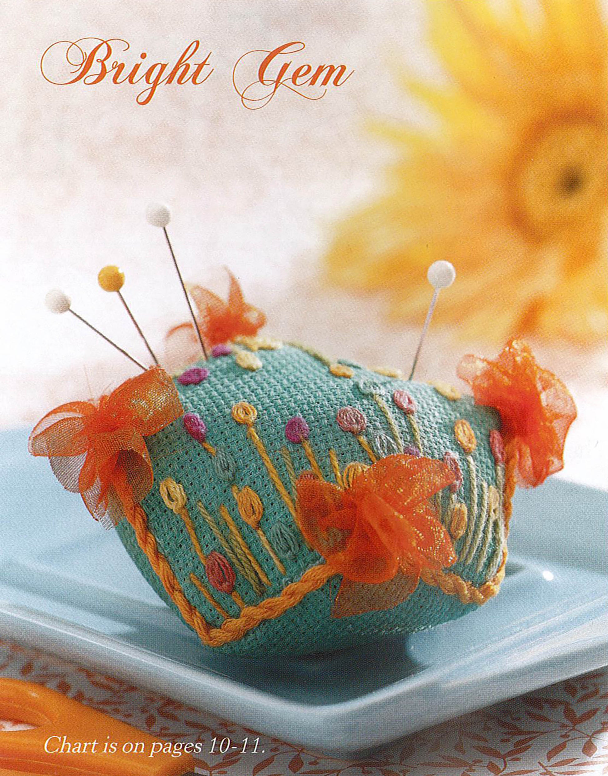 The Leisure Arts "Teach Yourself to Make Biscornu" digital download shows a decorative flower-embroidered pincushion with orange ribbon bows and pin heads, displayed on a blue dish with a blurred yellow flower in the background.
