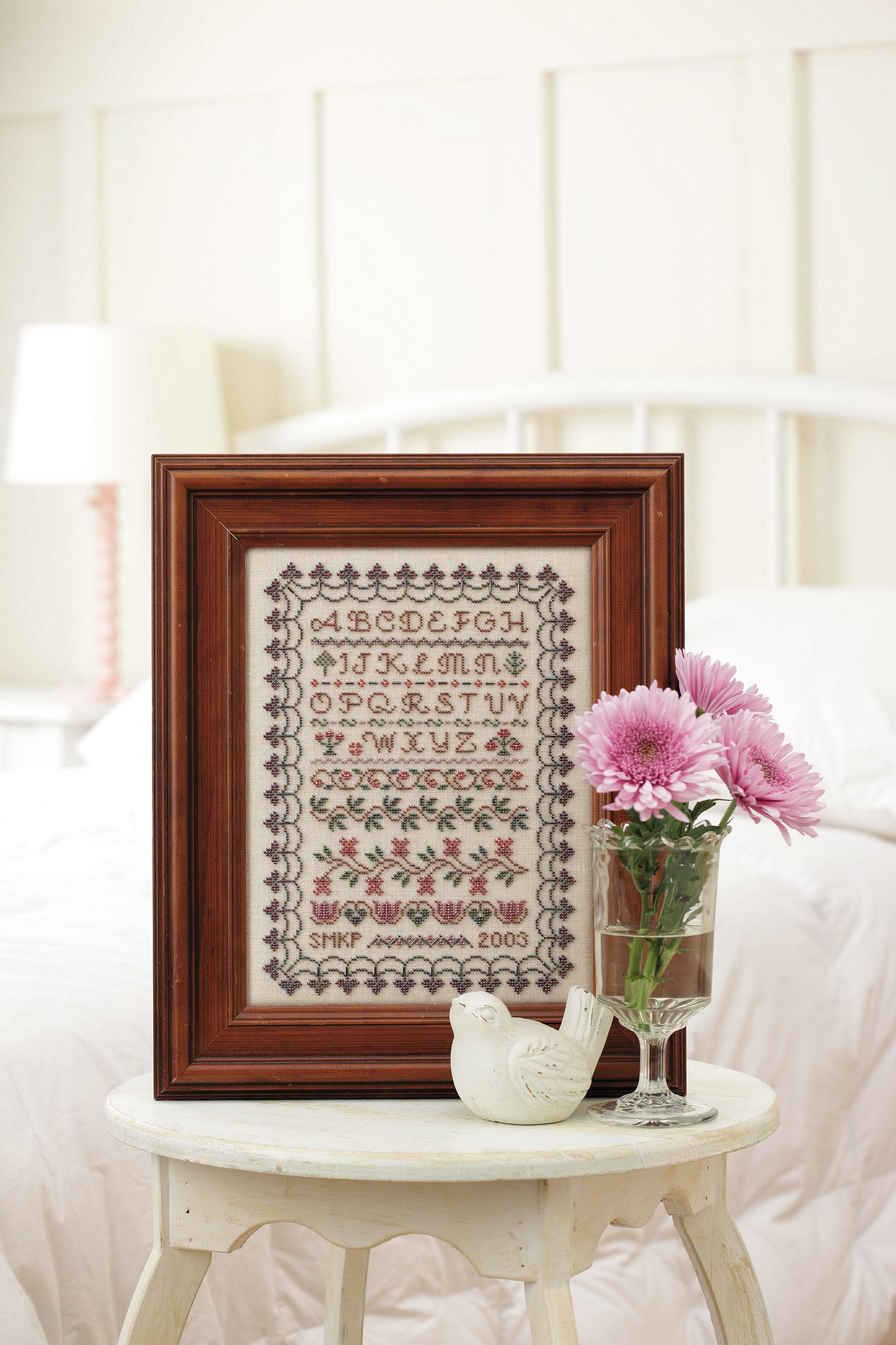 The Stitch a Beaded Garden—18 Designs Charted for Beading and Cross Stitch Digital Download by Leisure Arts is displayed framed on a bedside table with pink flowers, a ceramic bird, and a made bed in the background.